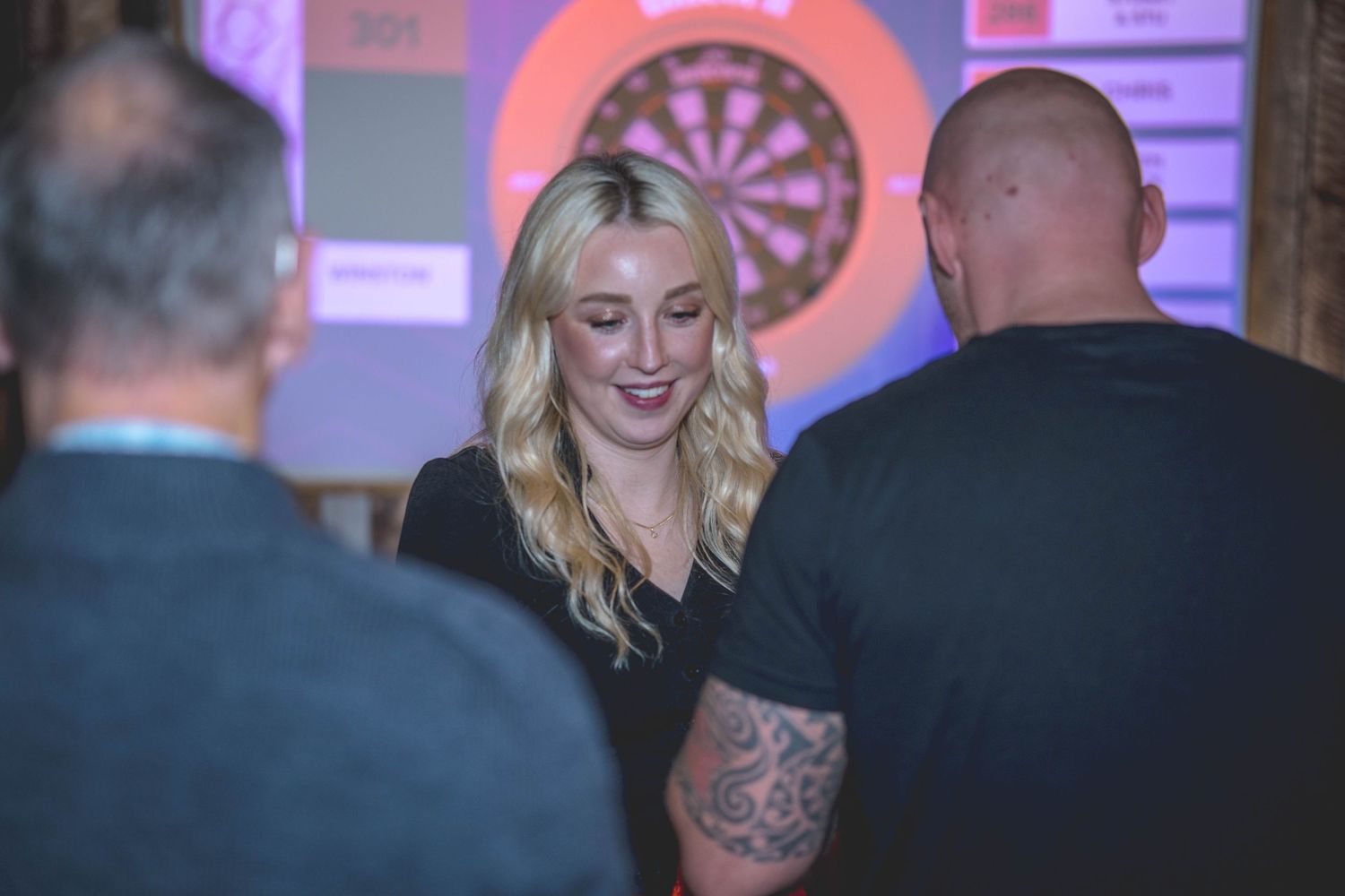 Blonde woman in black top smiling at a darts event, with a dartboard visible in the background.