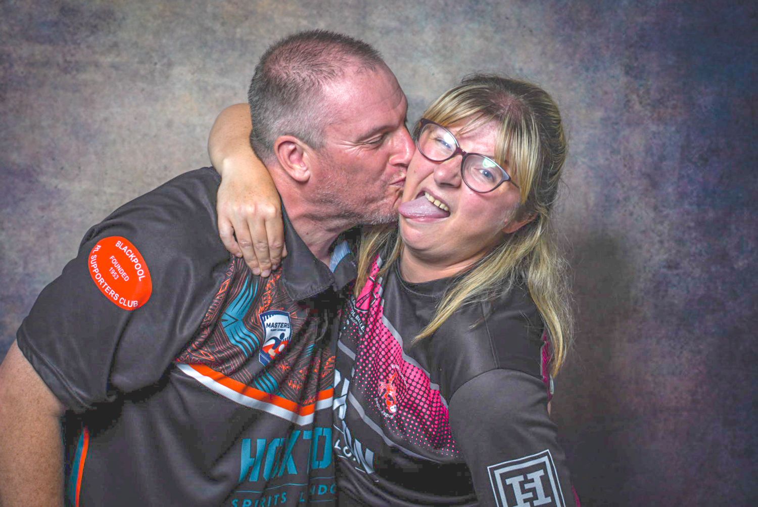 A man kisses a smiling woman on the cheek as she laughs, both wearing casual shirts against a dark background.