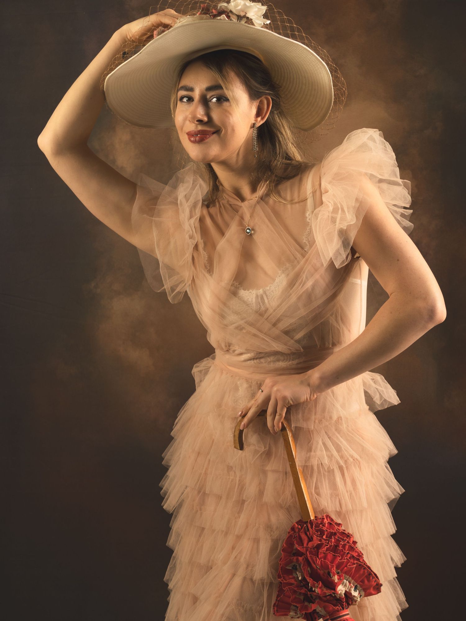 Woman in ruffled blush tulle dress and wide-brim hat holding a red floral umbrella, posing elegantly.