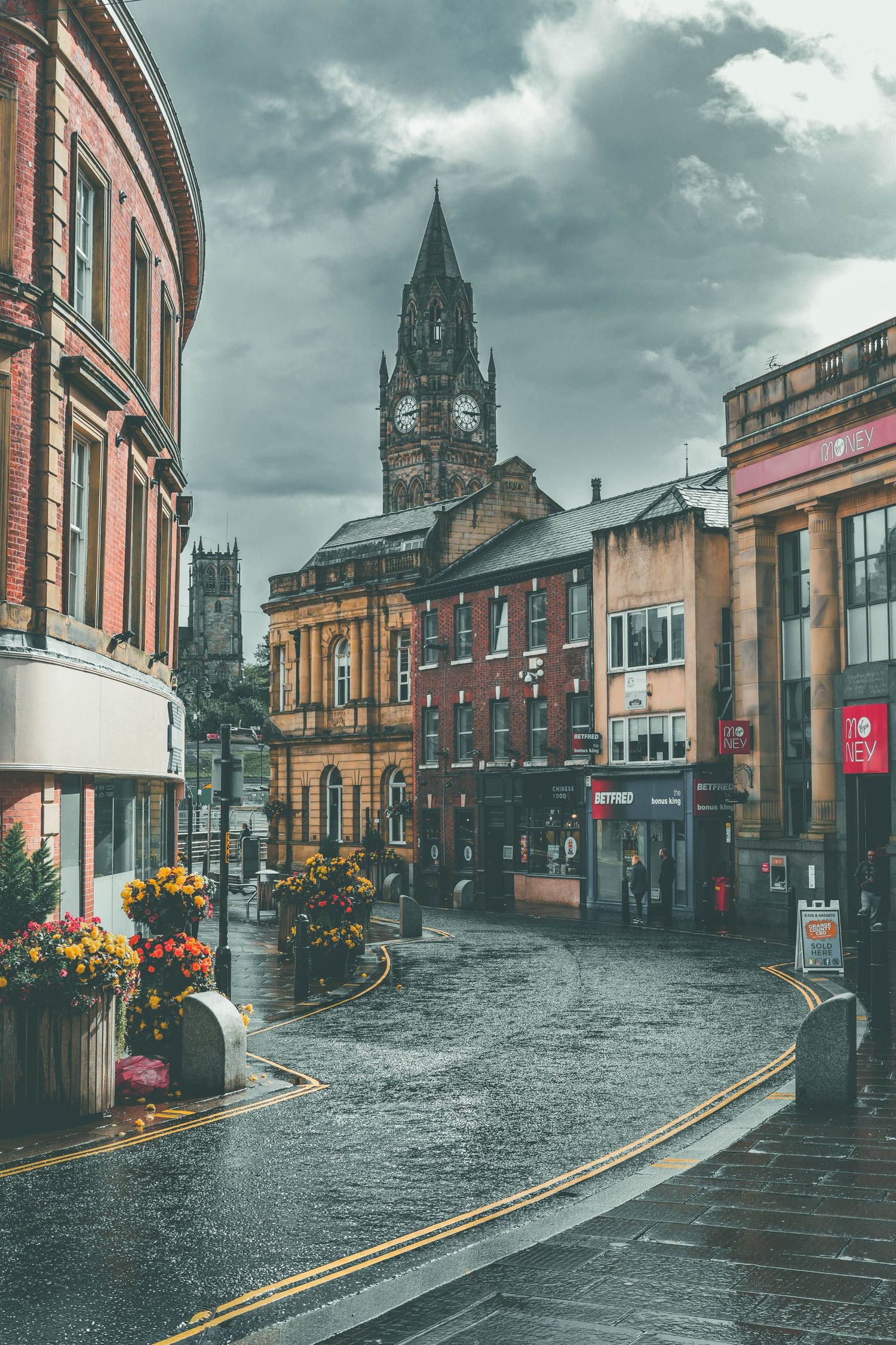 Historic cobblestone street in Rochdale with Victorian buildings and church spires under moody cloudy skies.