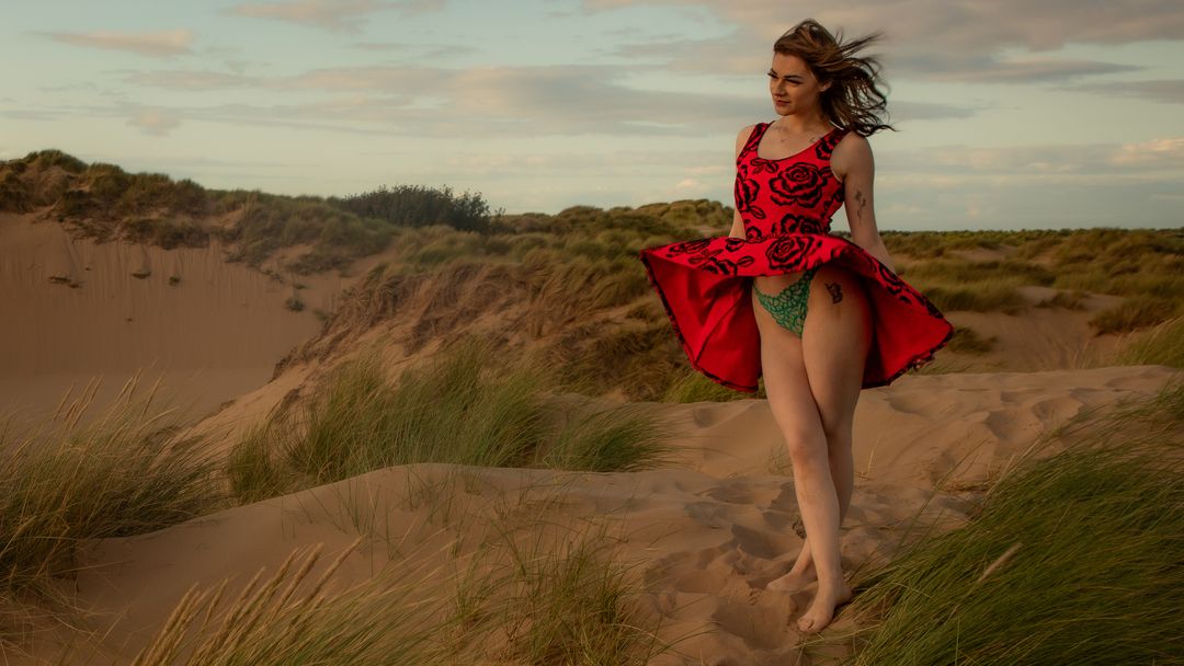 Woman in red dress walking on sandy dunes with green grass, wind blowing her skirt in a dramatic outdoor scene.