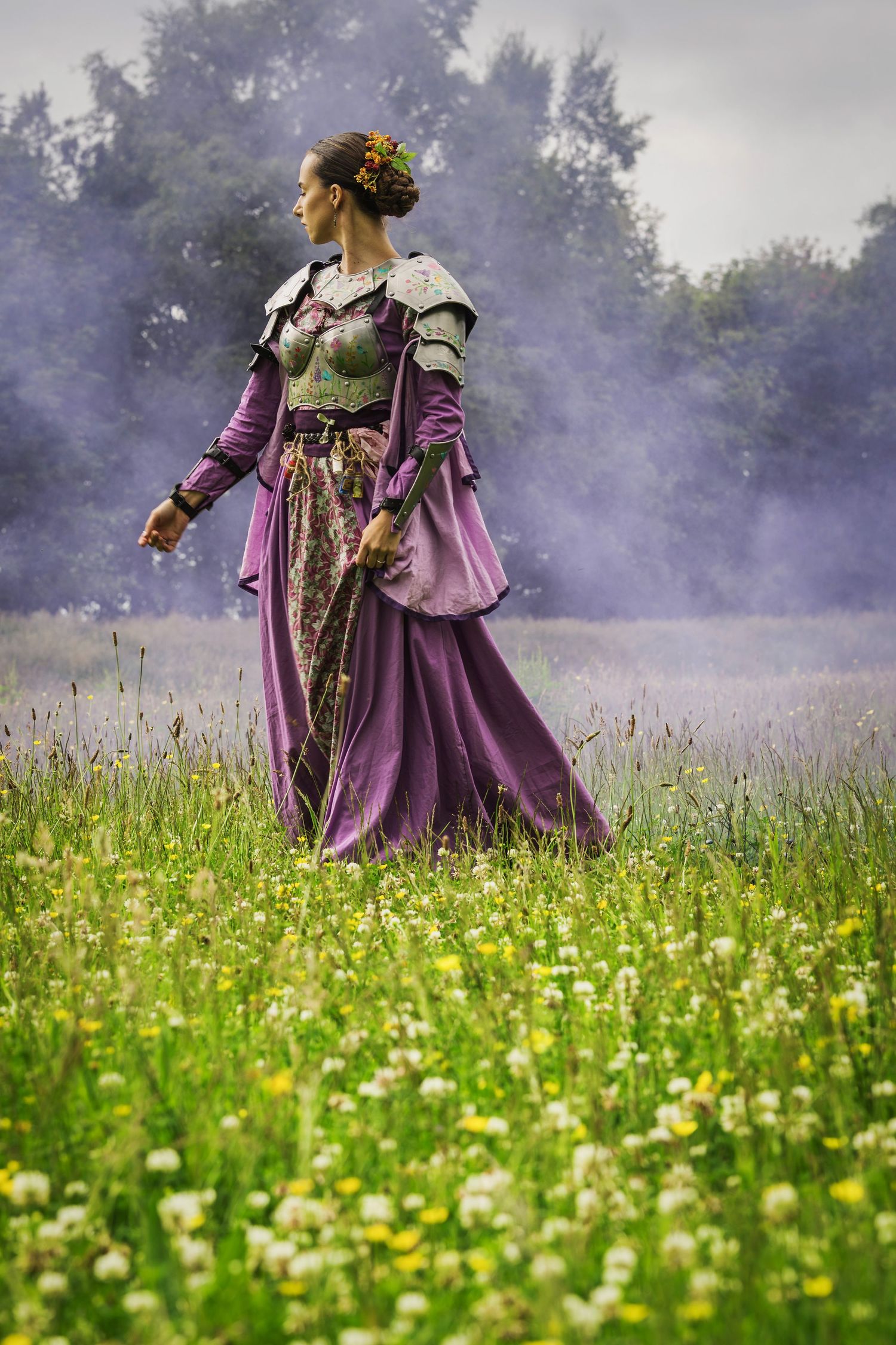 Woman in purple medieval gown with armor walks through a misty wildflower meadow with trees in the background.