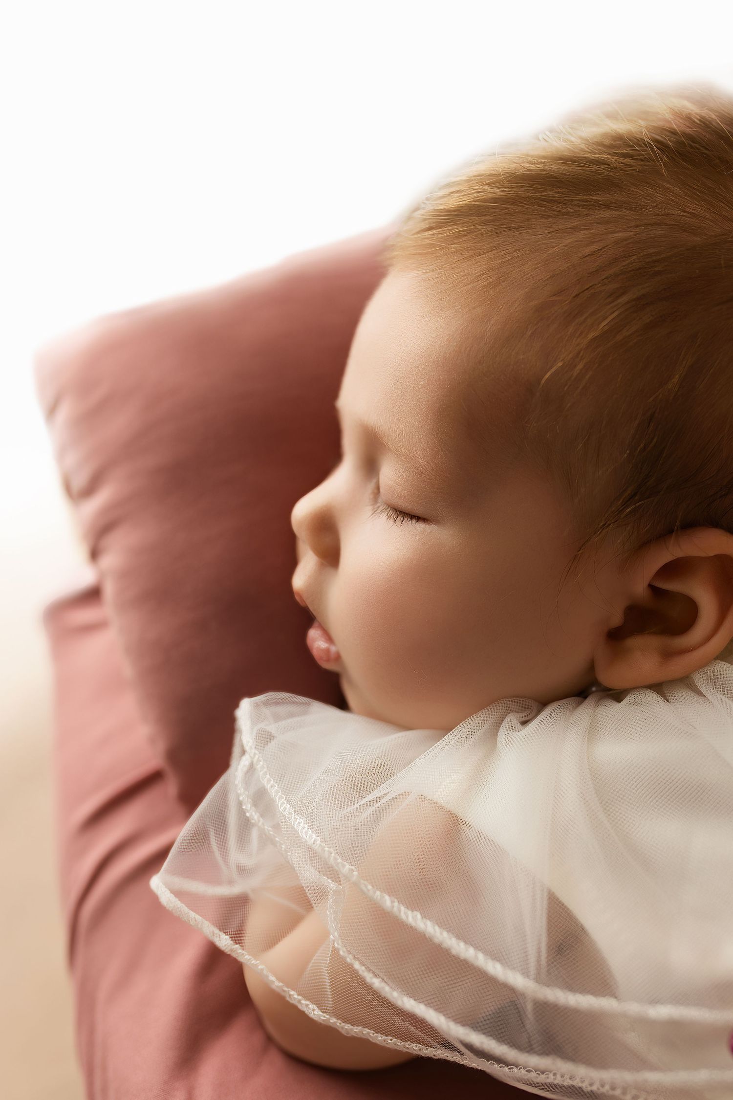 Sleeping newborn baby in delicate white lace wrap against pink background.