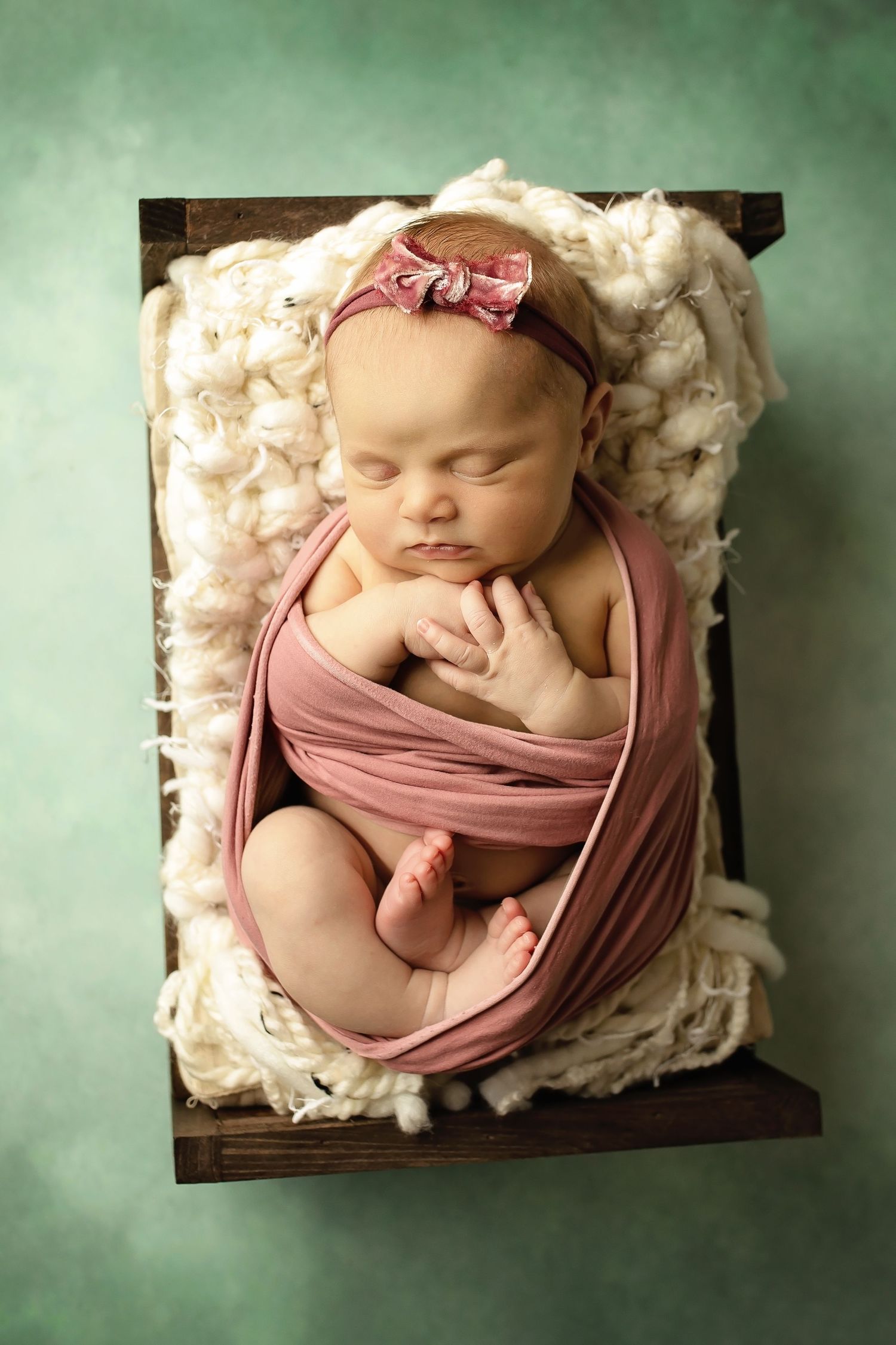 Newborn baby sleeping peacefully in a wooden box lined with white wool and wrapped in dusty rose fabric.