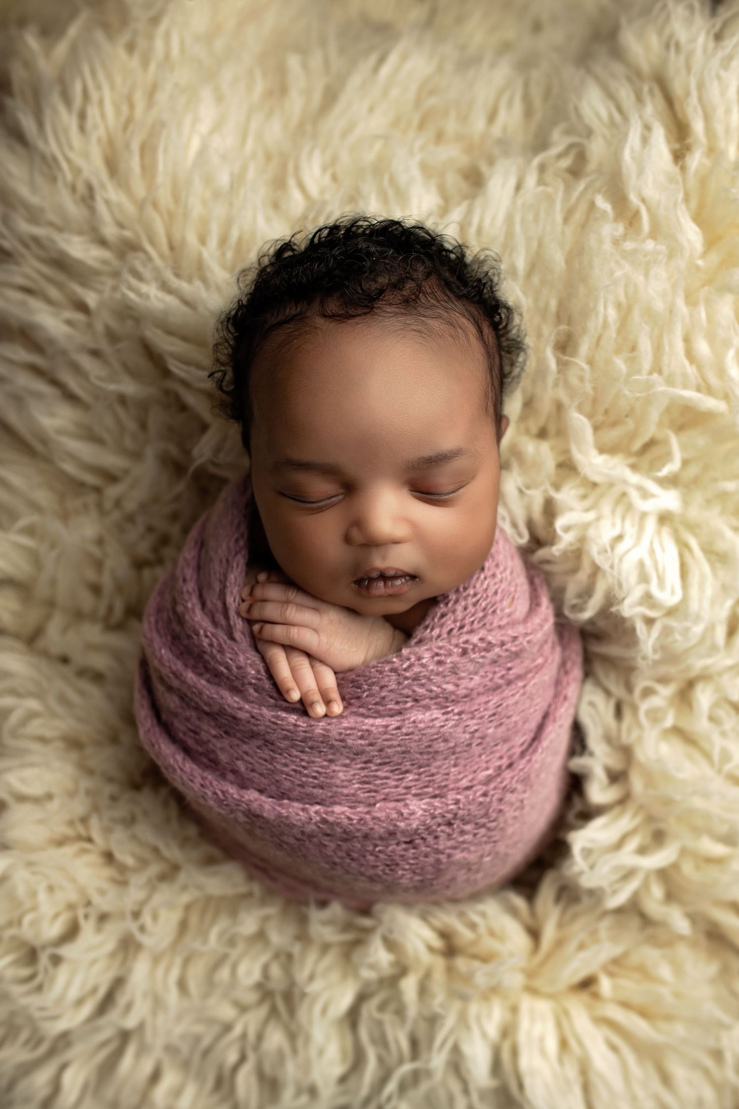 Professional newborn photos of baby wrapped in pink knit fabric on cream-colored fur backdrop.