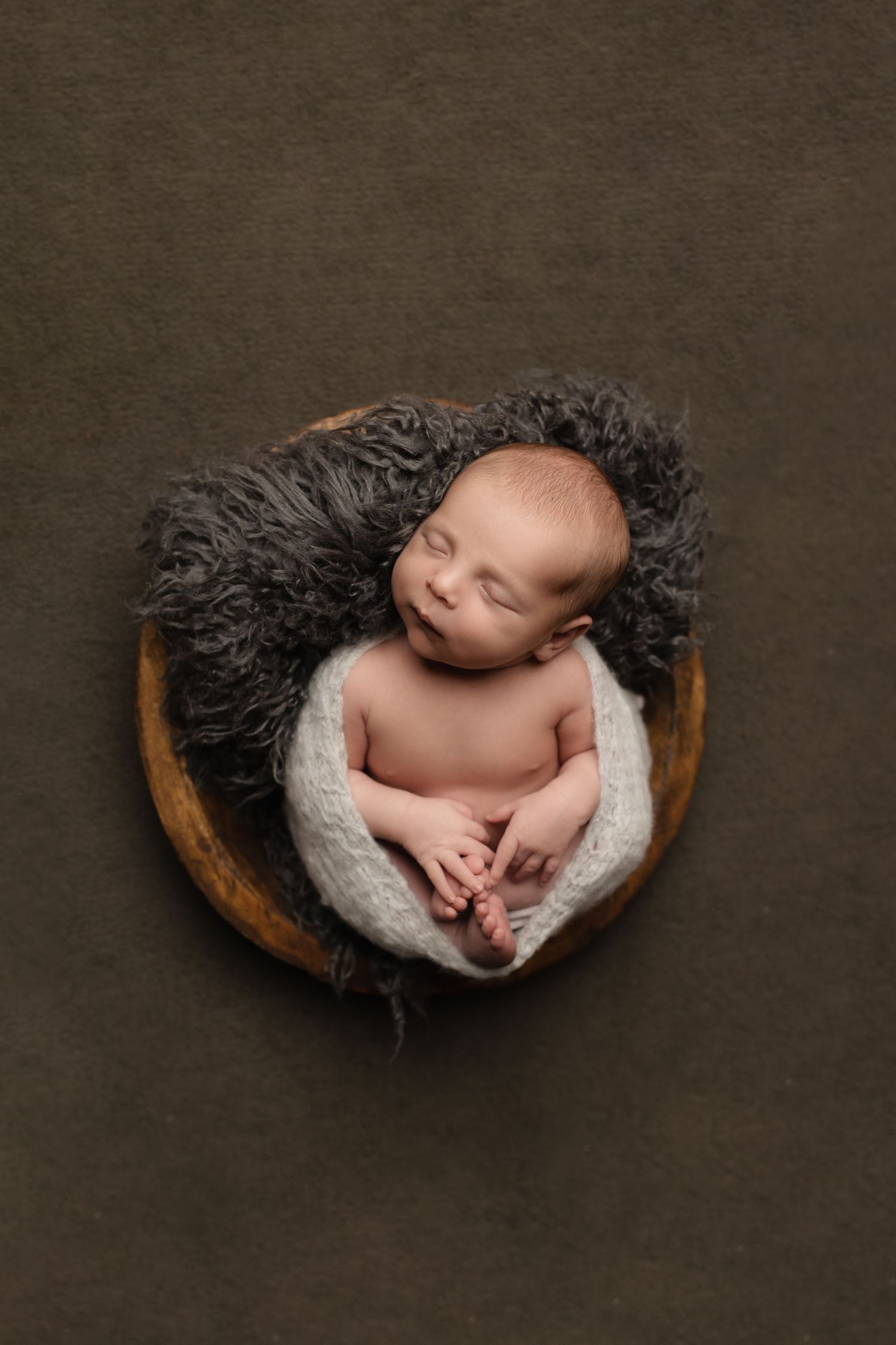 Artistic newborn photos showing baby sleeping in wooden bowl with dark textured fabric.