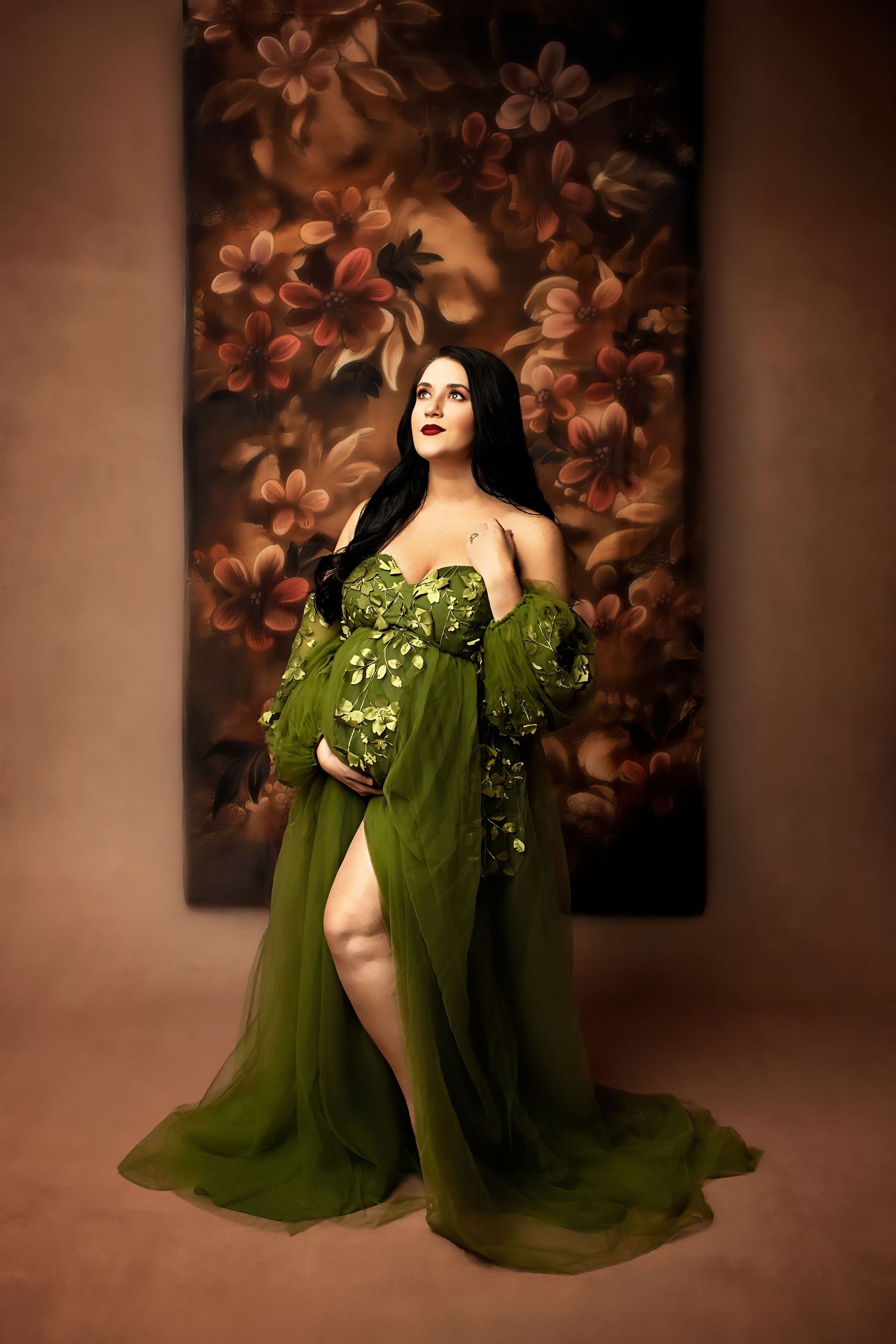 Elegant olive green gown with flowing sheer fabric and floral embellishments against a warm brown floral backdrop.