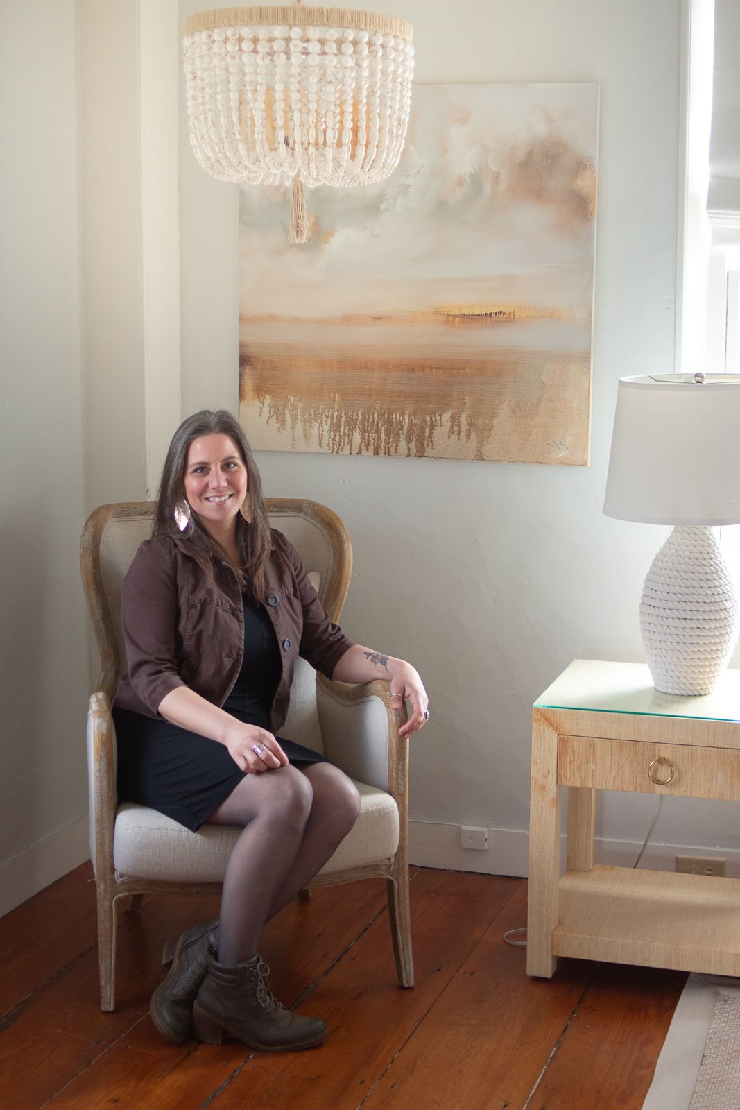 Seated figure in a brown jacket and boots next to beaded chandelier and coastal artwork in cozy room setting.