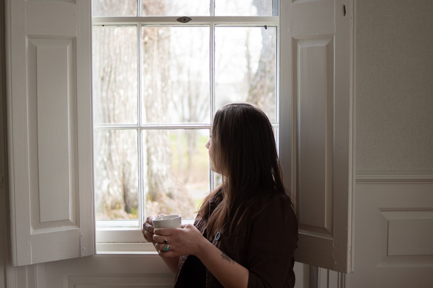 Silhouette of someone holding a cup while looking out an open window with white shutters.