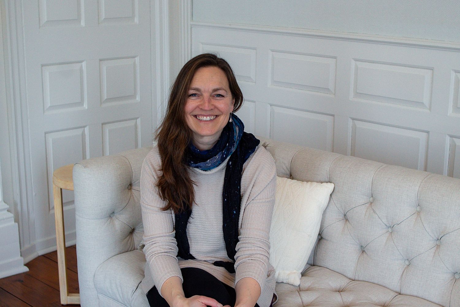 Person in a cream sweater and navy scarf sits smiling on a white tufted couch with decorative pillows.