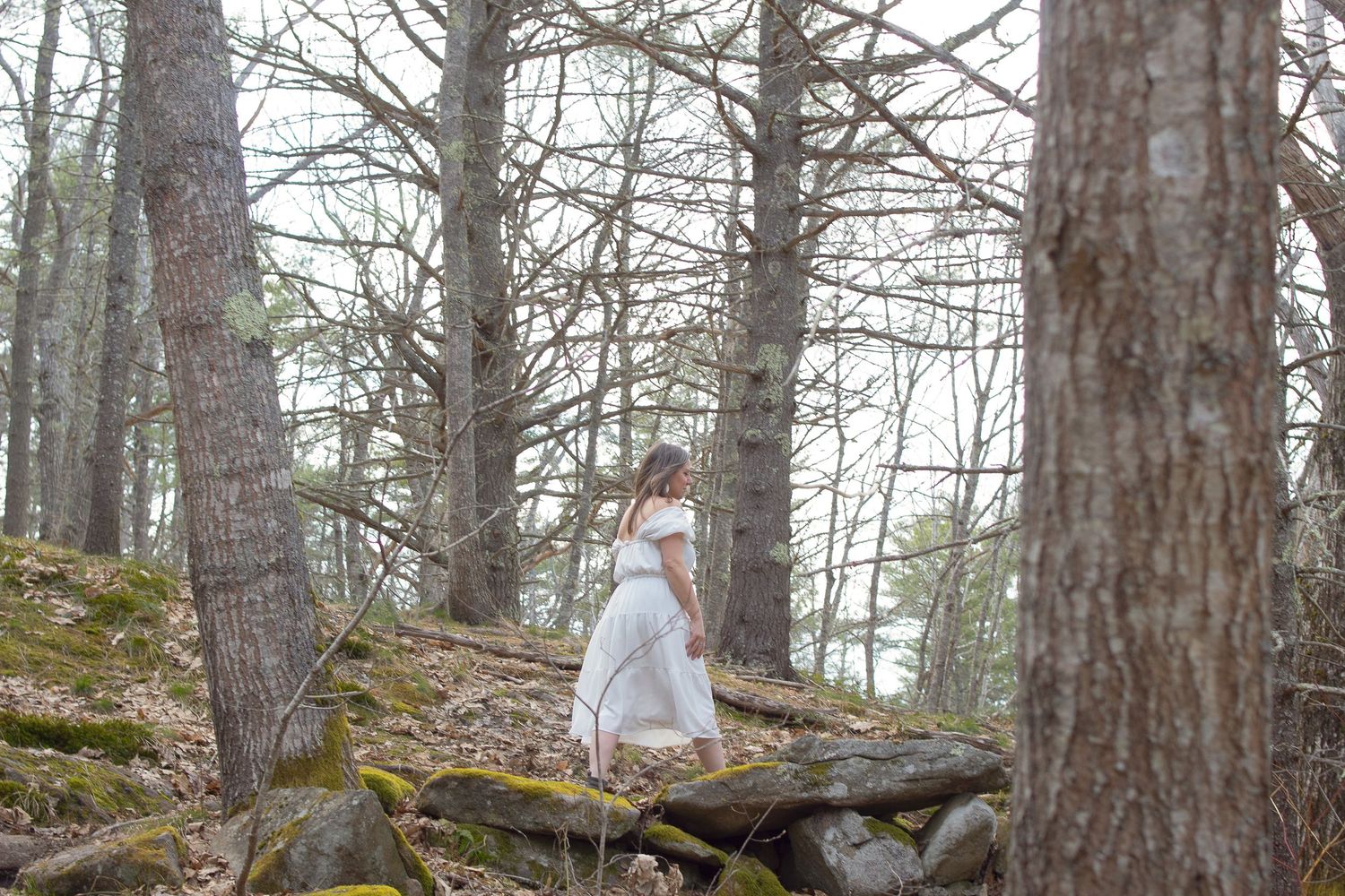 A figure in a flowing white dress stands gracefully on fallen logs in a misty forest setting.