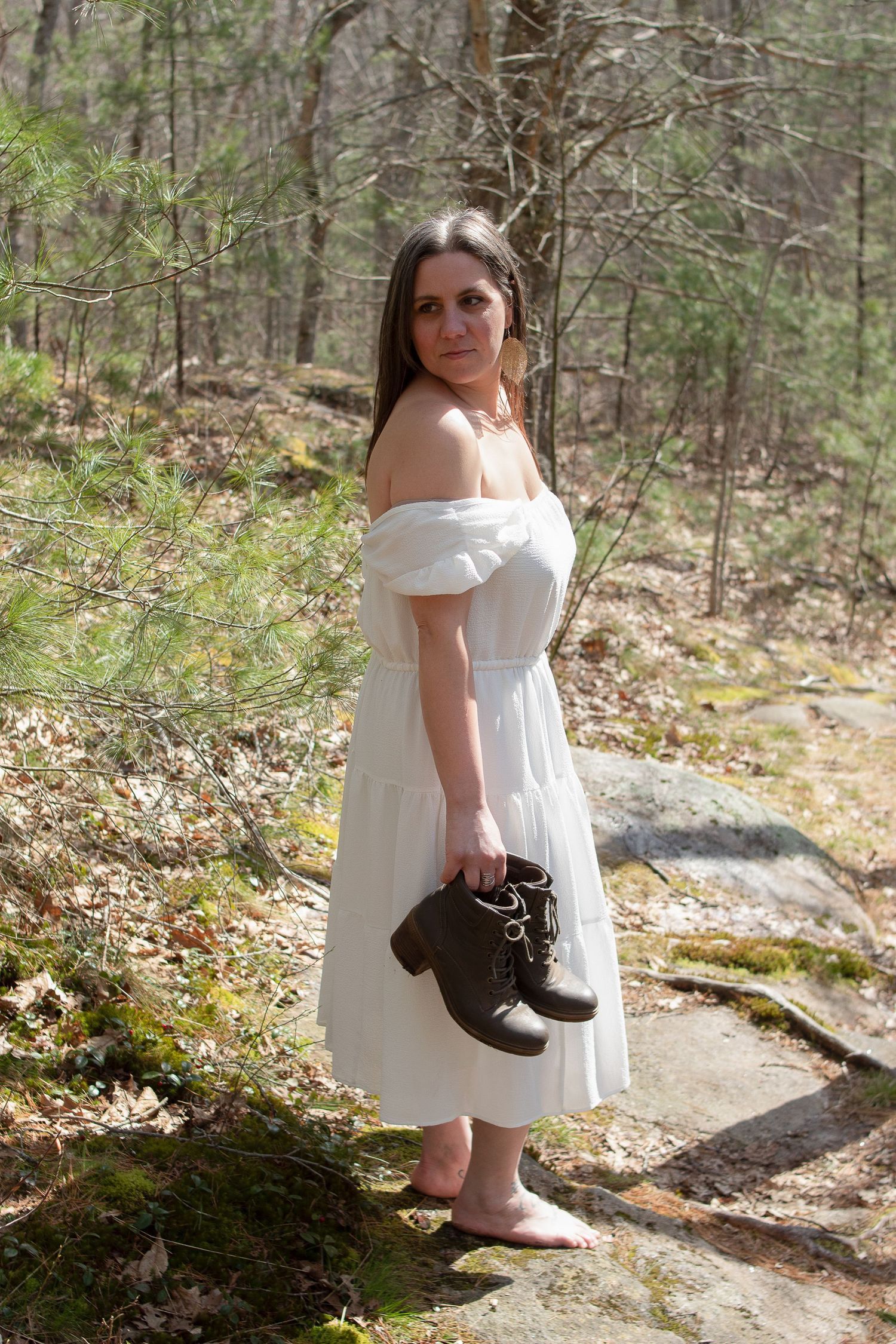 A person in a white dress holds brown boots while standing on moss-covered rocks in the forest.