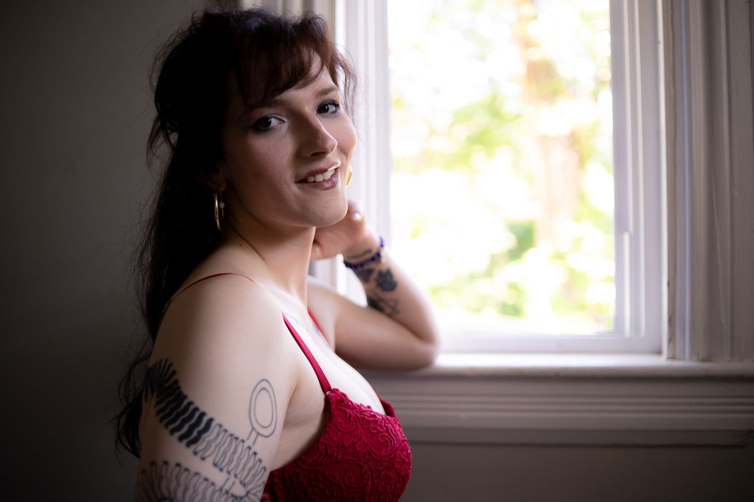 A woman in a red dress poses by a window in a romantic boudoir photography session.