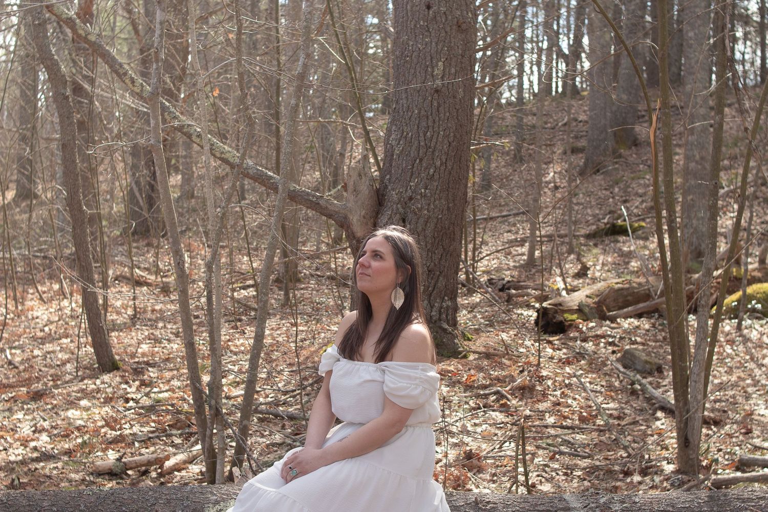 A person in a white dress sits elegantly in a forest clearing surrounded by bare trees and fallen leaves.
