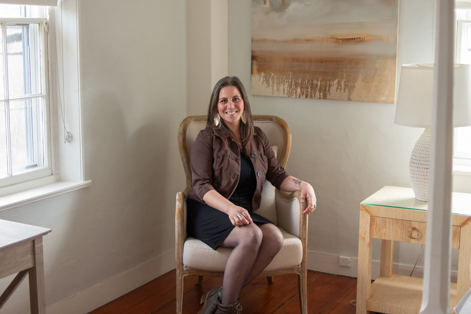 Someone sits cross-legged in an armchair by a window in a bright room with neutral wall art.