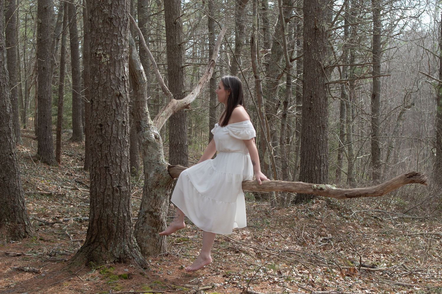 A person in a white dress performs a graceful dance pose among tall forest trees.