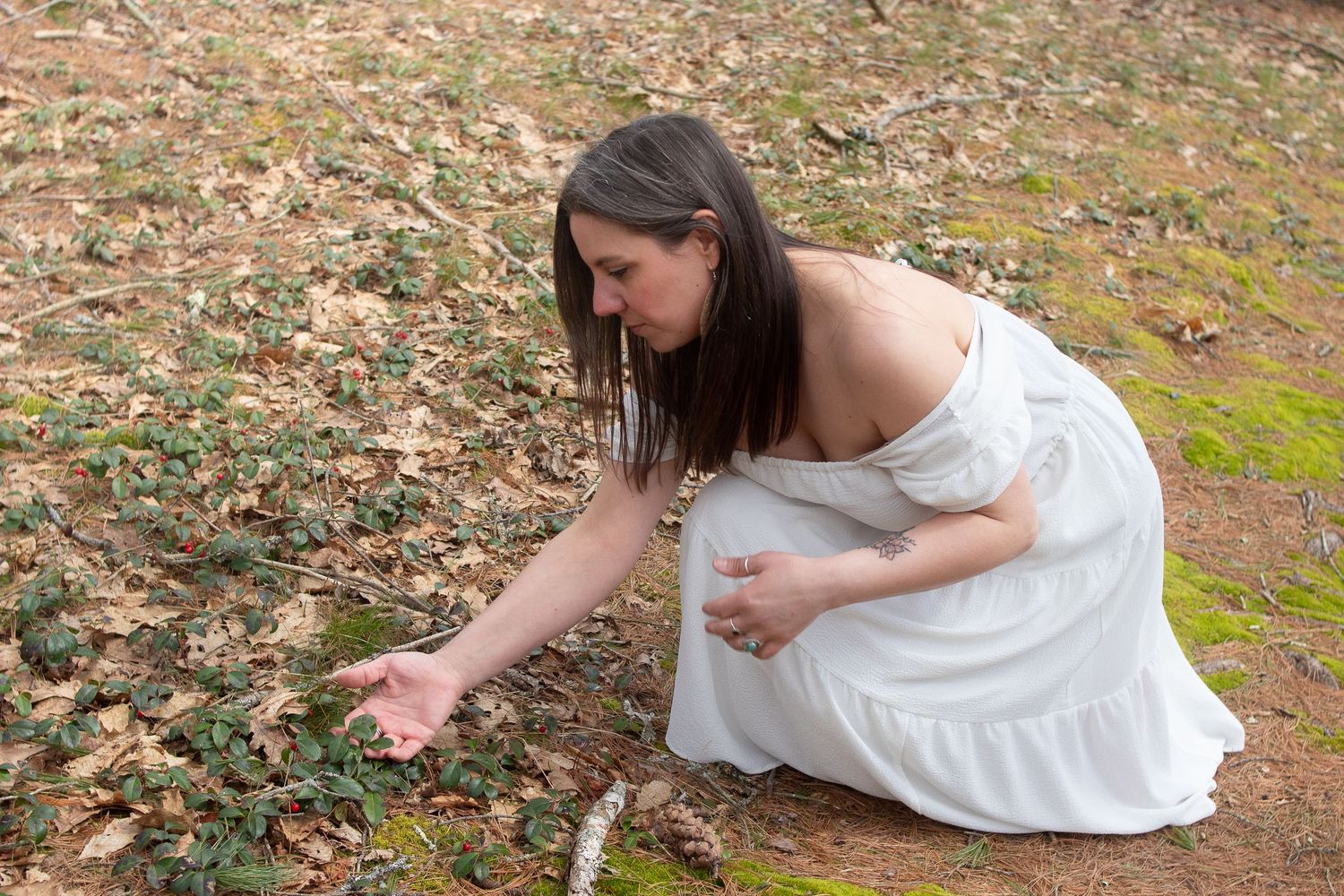 A person in a white dress reaches down to touch fallen leaves on mossy ground.