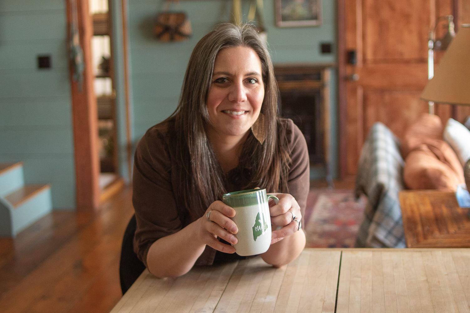 Someone holds a warm beverage while leaning on a rustic wooden table in a cozy home interior setting.
