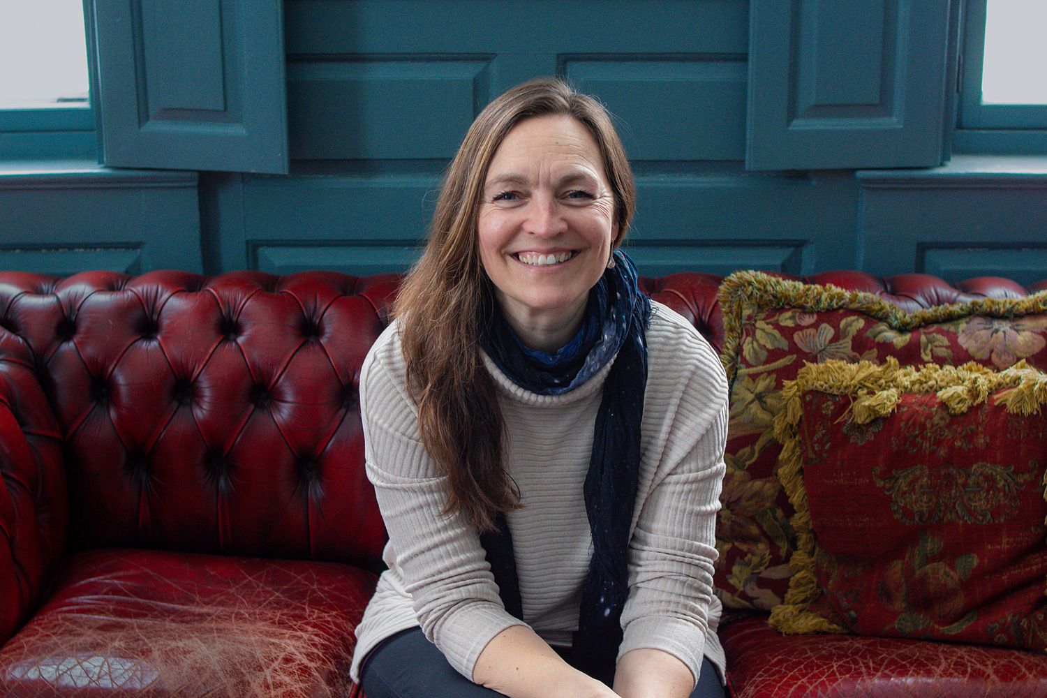 A person sits smiling on a burgundy tufted leather couch with decorative throw pillows against a teal wall.