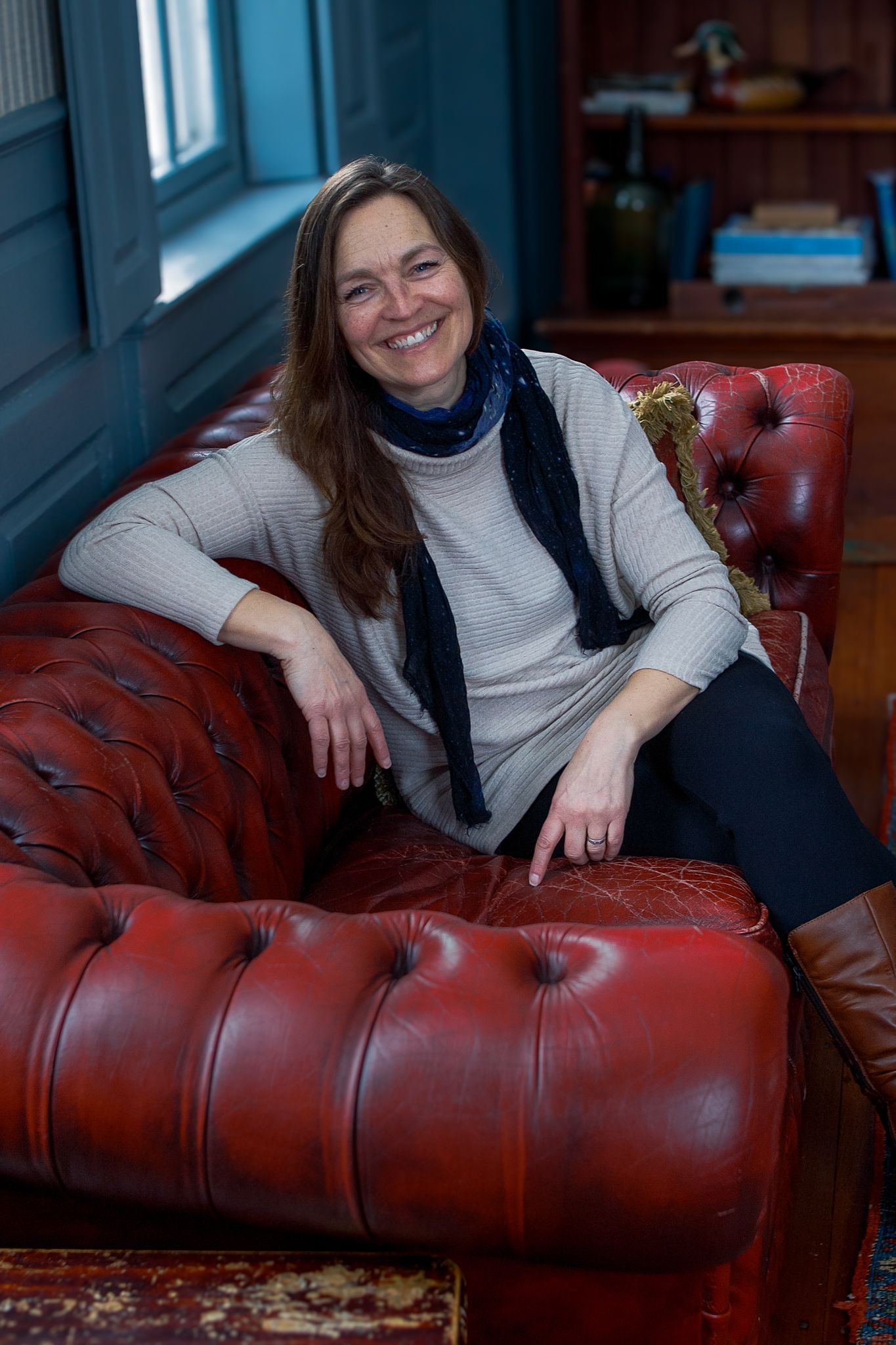 Person relaxing on a red leather Chesterfield sofa near a window in a cozy room.