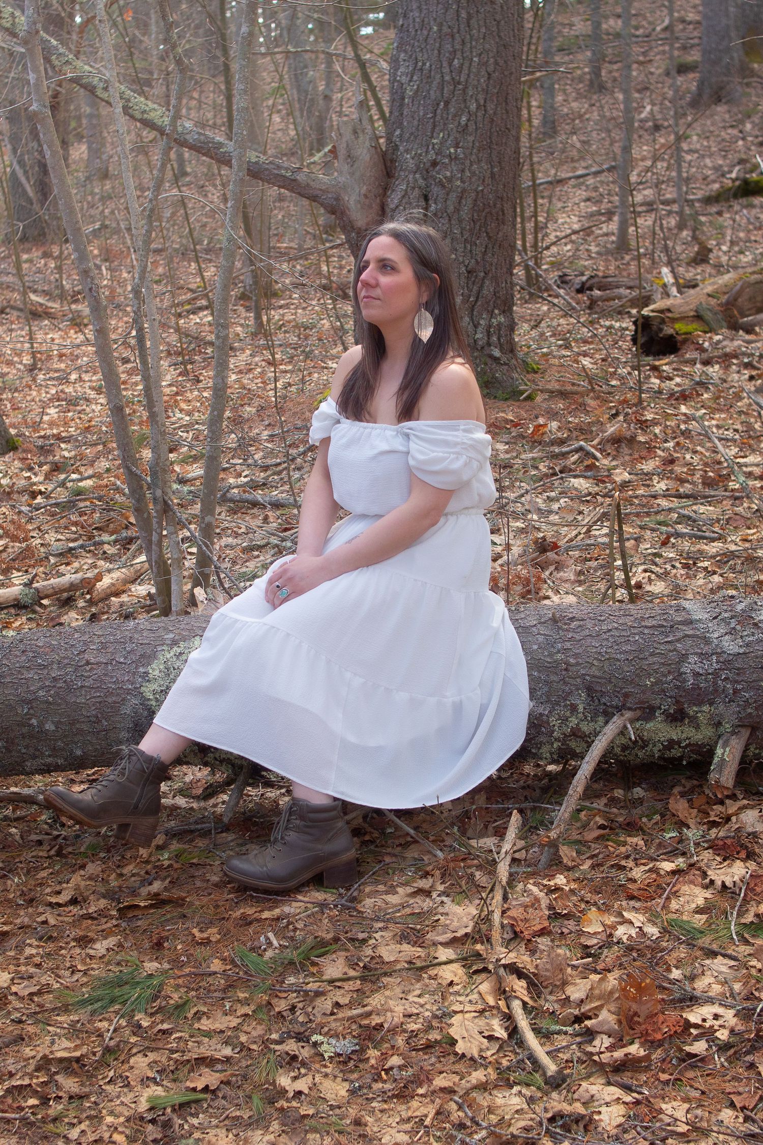 A person in an off-shoulder white dress sits on a fallen log in an autumn forest.