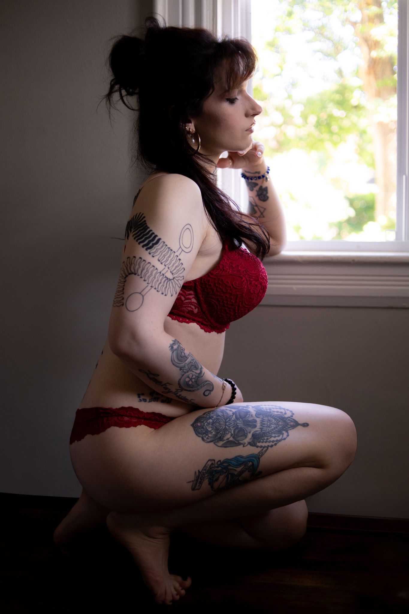 Artistic boudoir photograph showing a silhouetted figure with tattoos sitting near a window in red lingerie.