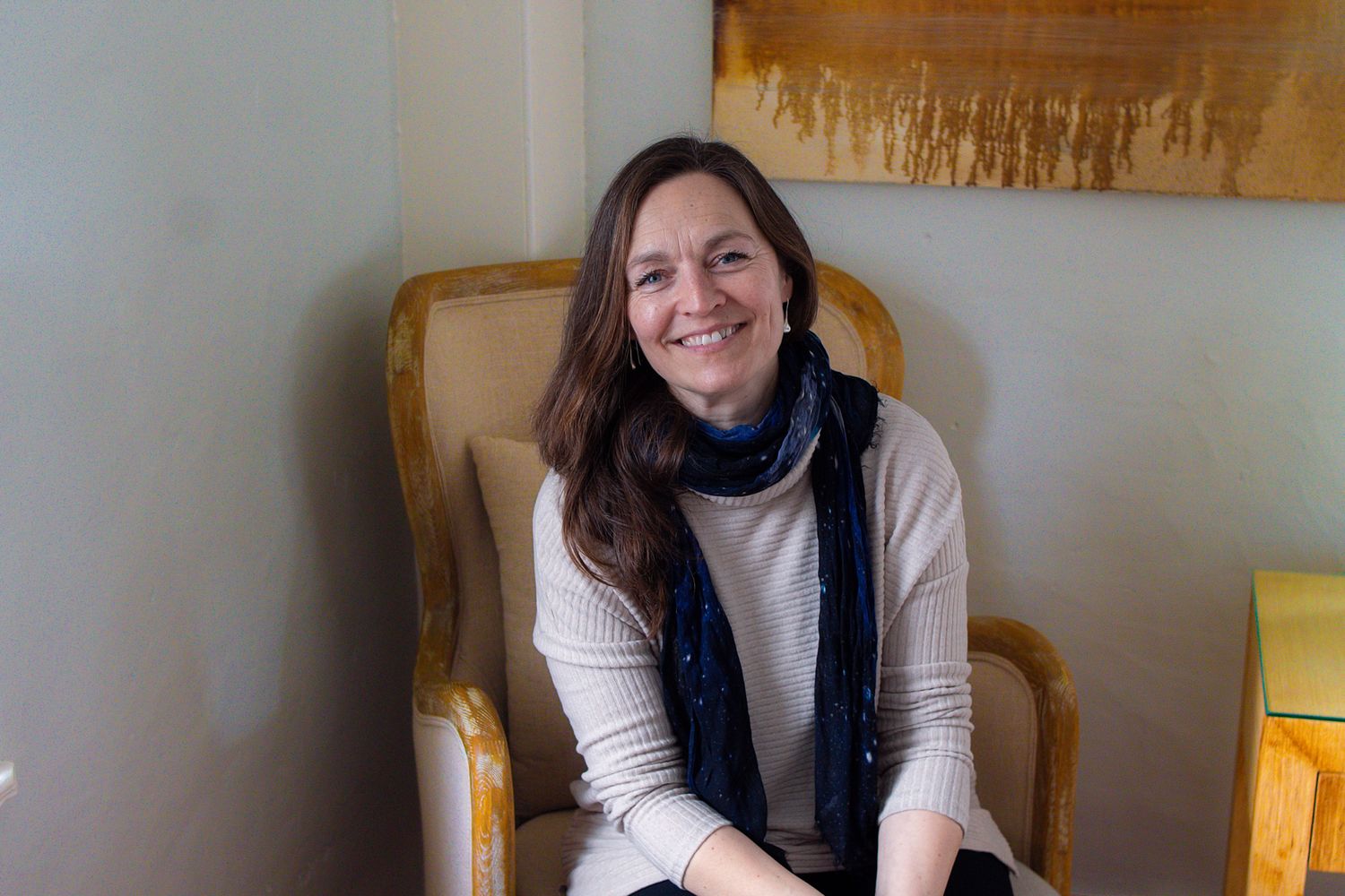 A person in a white sweater and dark scarf sits smiling warmly in a wooden chair against a beige wall.