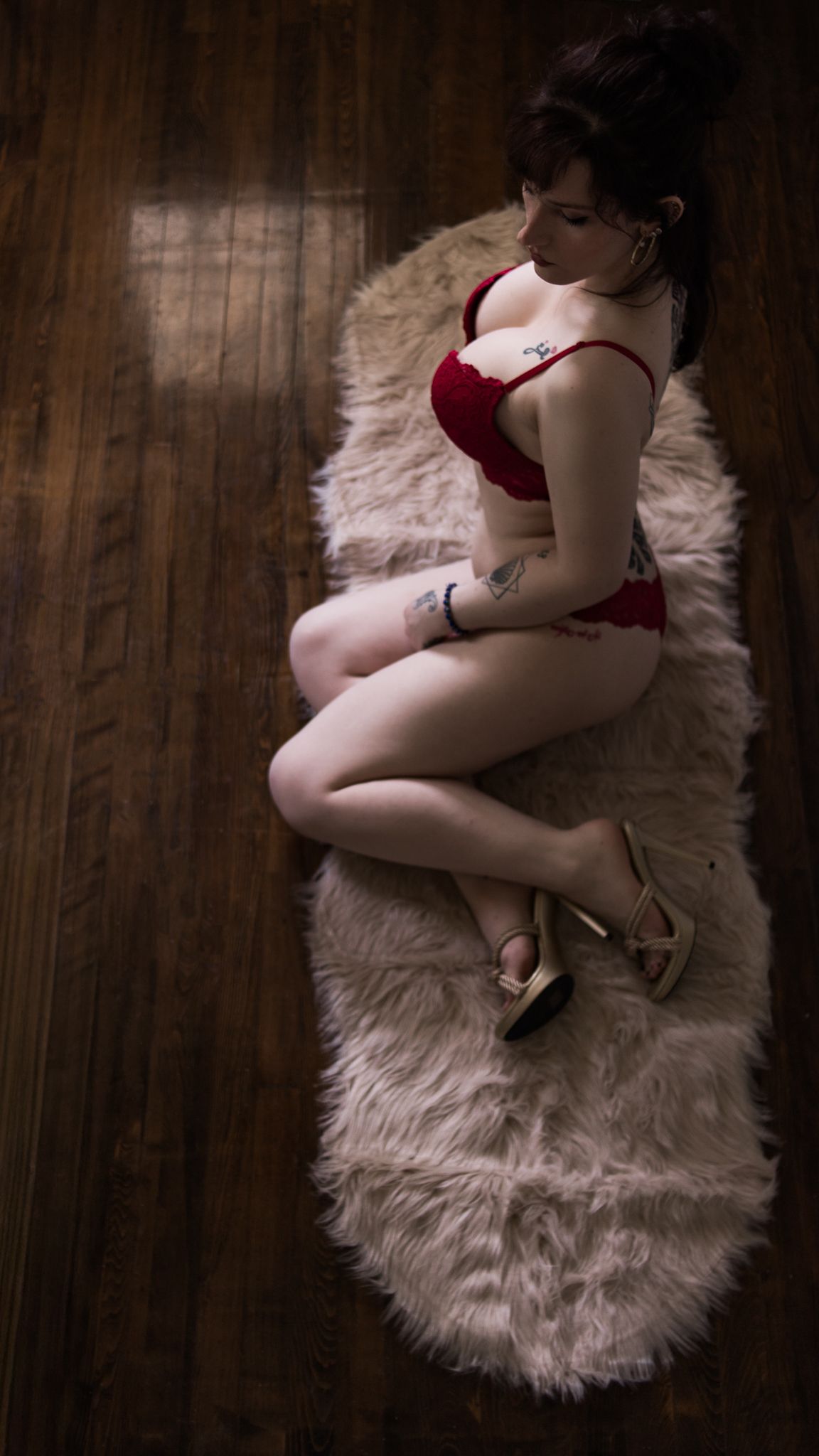 A figure in red lingerie reclines on a plush fur rug on hardwood flooring in moody boudoir lighting.