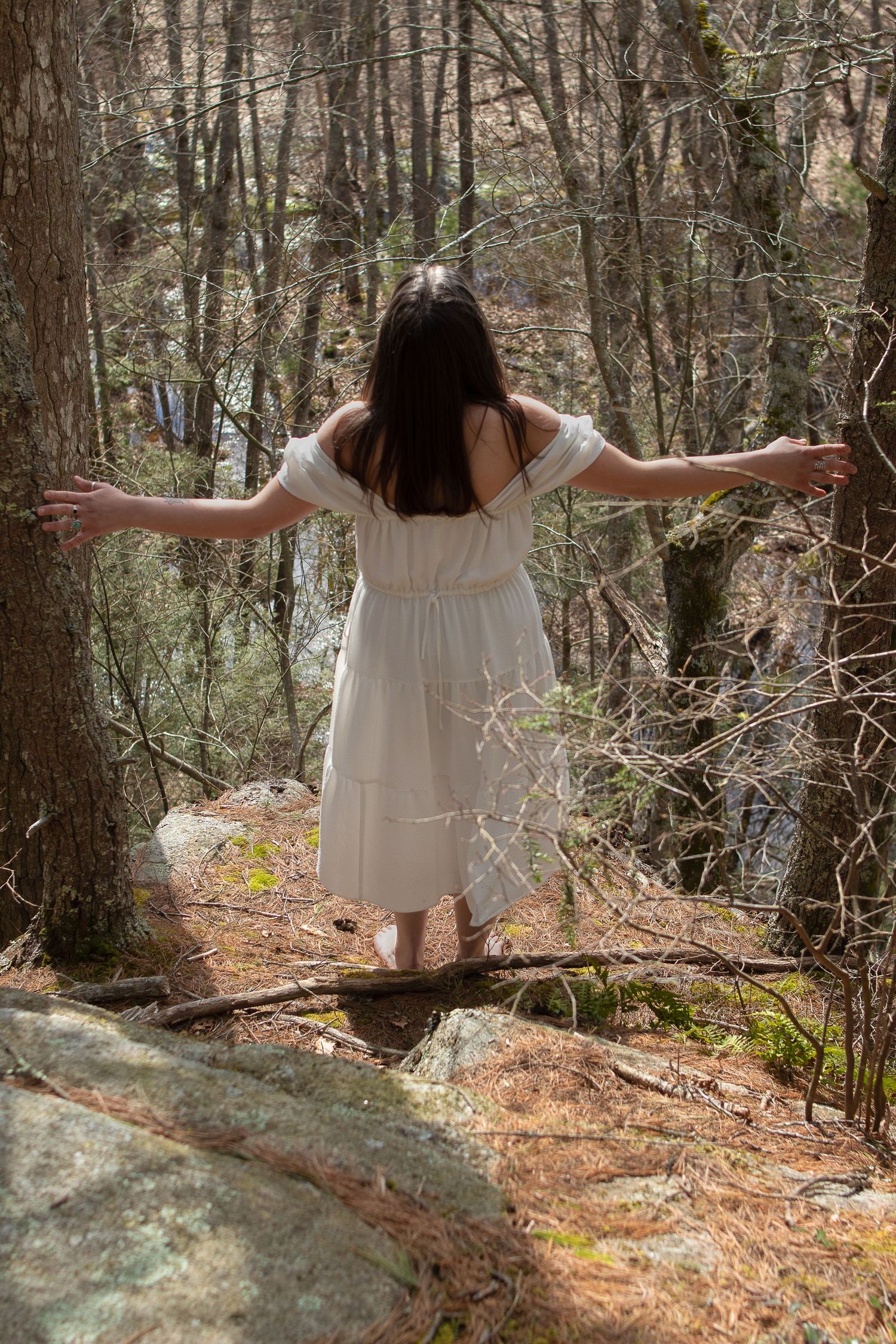 Person in flowing white dress stands with arms outstretched on rocky forest ledge.