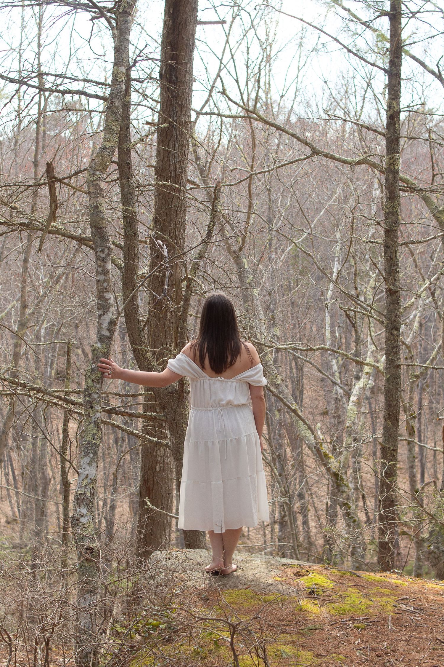 A person in a white dress stands on a mossy cliff edge overlooking a winter forest.