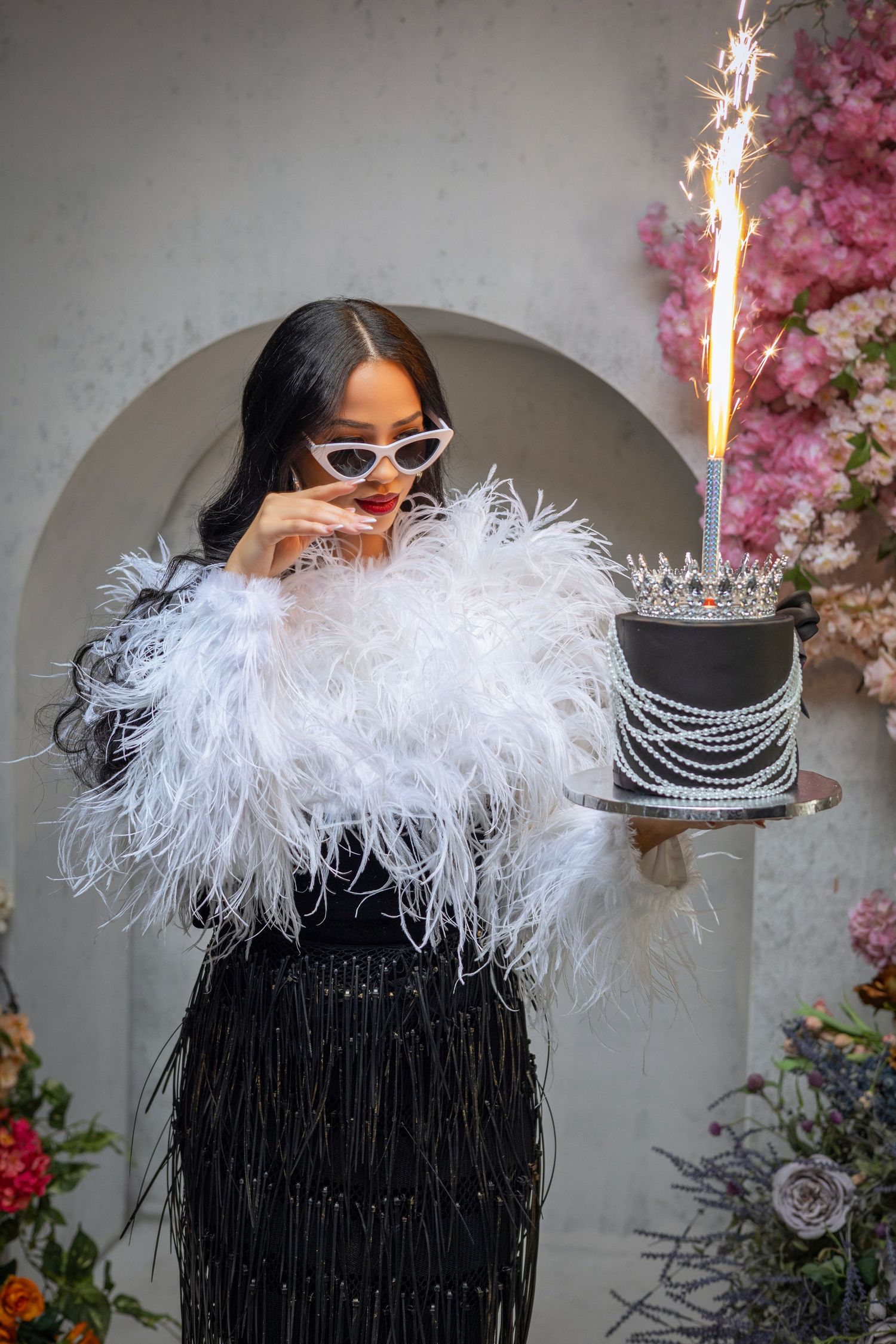 A glamorous party scene with a feathered white boa, sunglasses, black dress, and sparkler-topped birthday cake against floral decor.