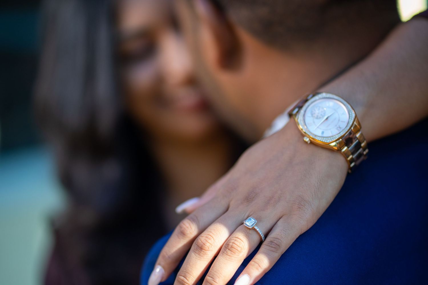 Closeup shot showing a gold wristwatch and engagement ring with soft bokeh lighting in the background.