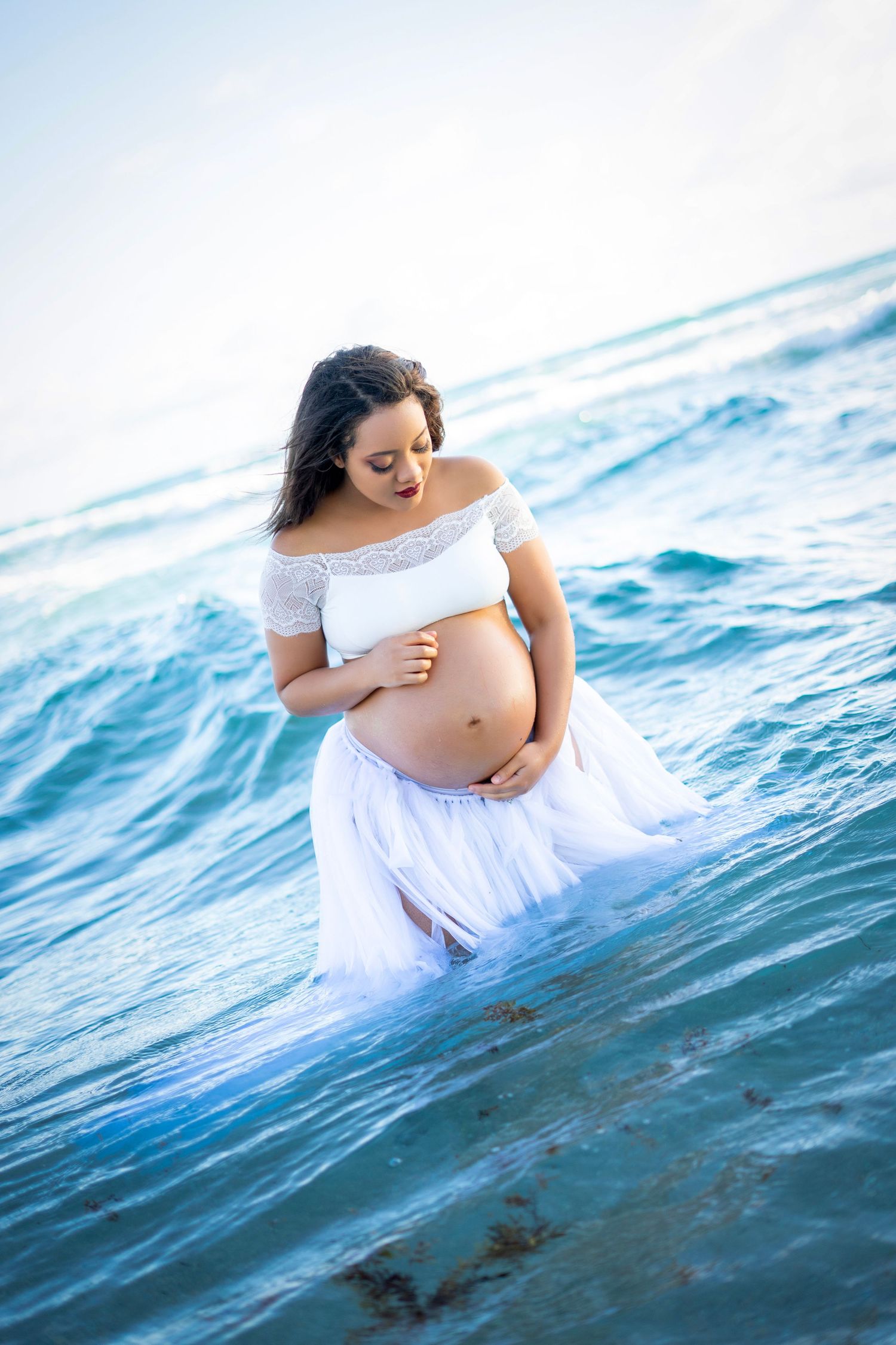 A maternity photoshoot in blue ocean waves with flowing white fabric creating an ethereal underwater effect.