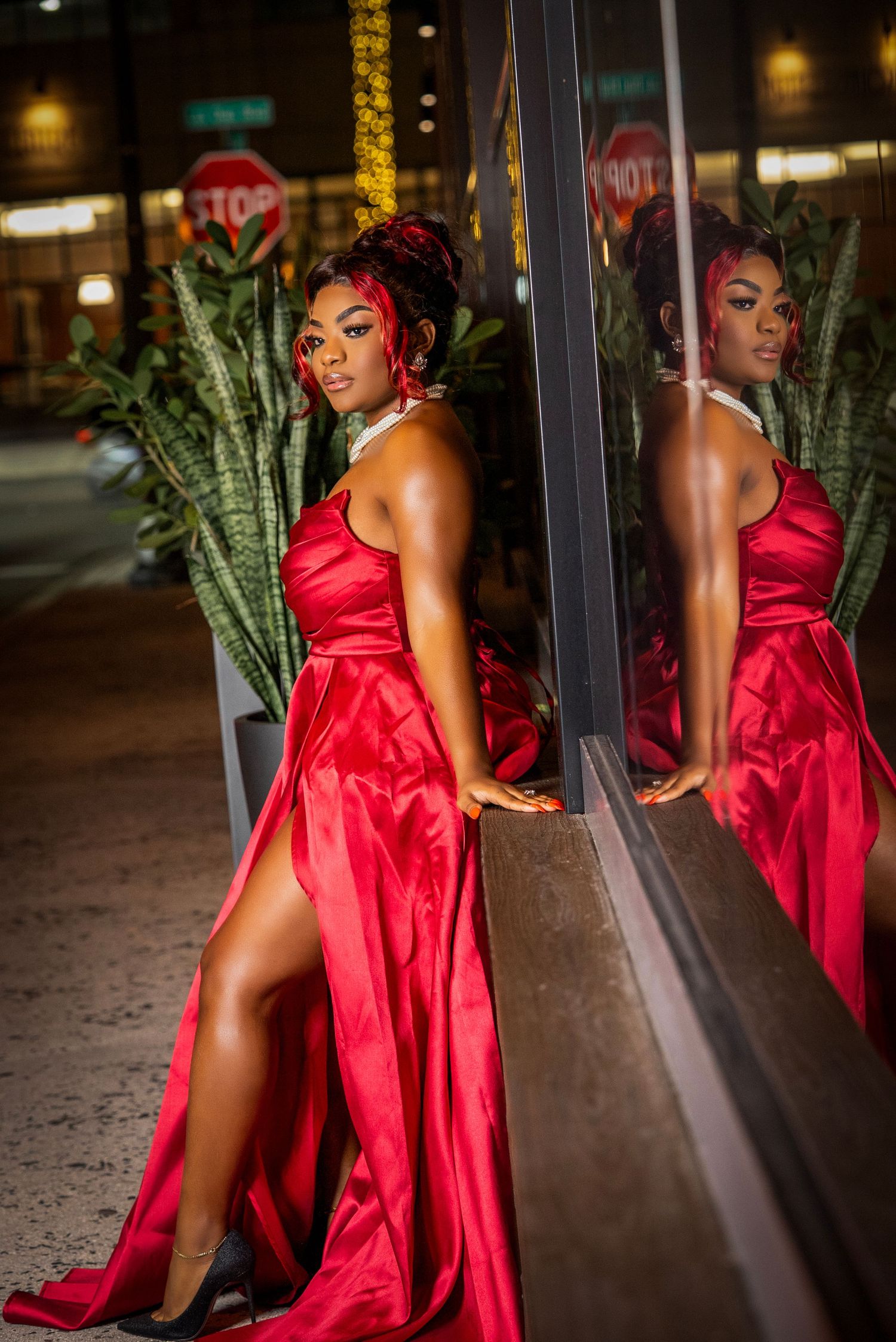 A glamorous evening scene featuring a flowing red satin gown reflected in a window at night with palm plants visible.