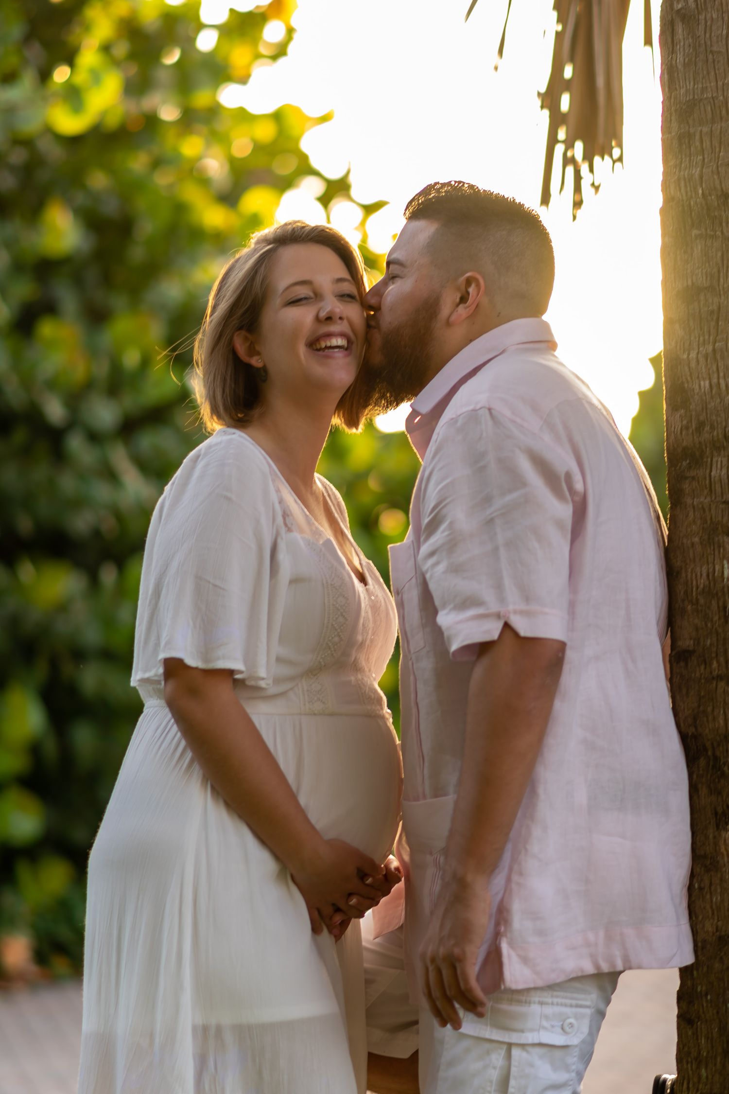 A couple shares a joyful moment at sunset as they embrace in white and pink attire against a golden backlit background.