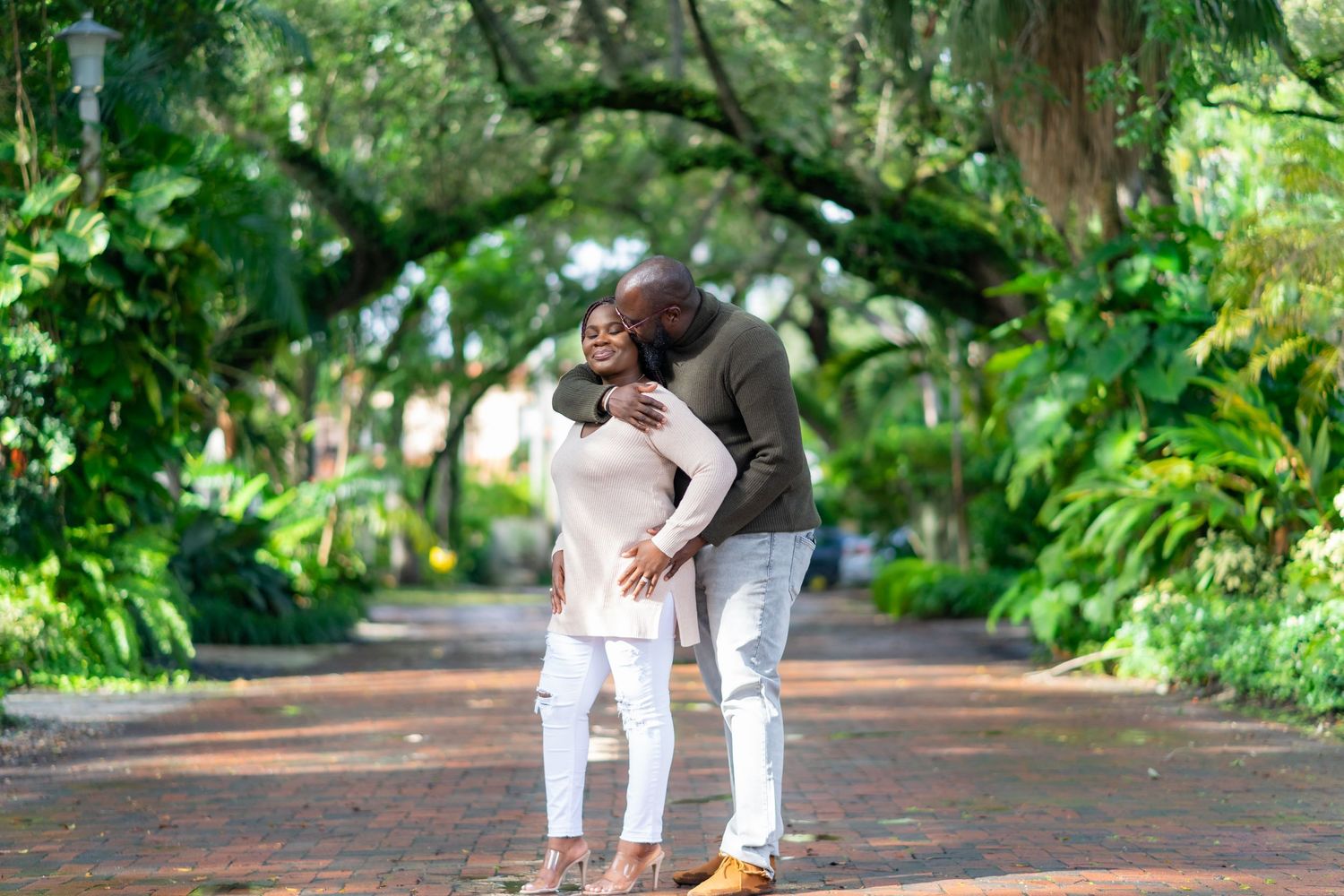 A couple embraces while standing on a tree-lined pathway in a lush garden setting.