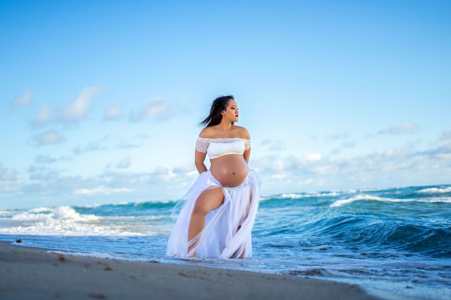 A serene maternity photo at the beach with crashing waves and blue sky in the background.