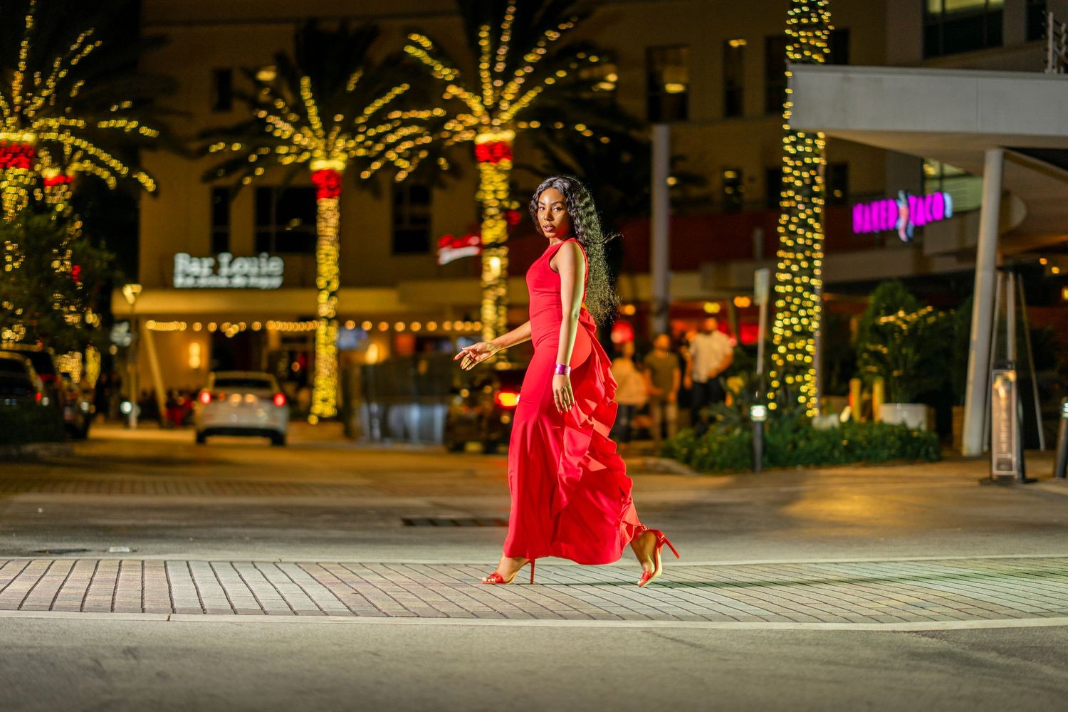 A person in a flowing red dress spins elegantly in an illuminated city plaza at nighttime with twinkling lights.