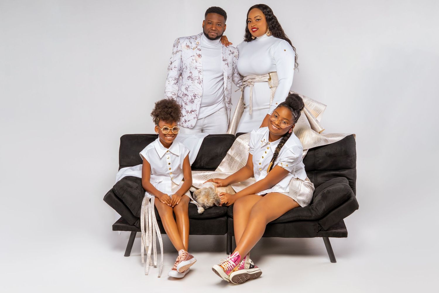 A family of four wearing coordinated white outfits poses together on a black modern sofa against a light gray background.