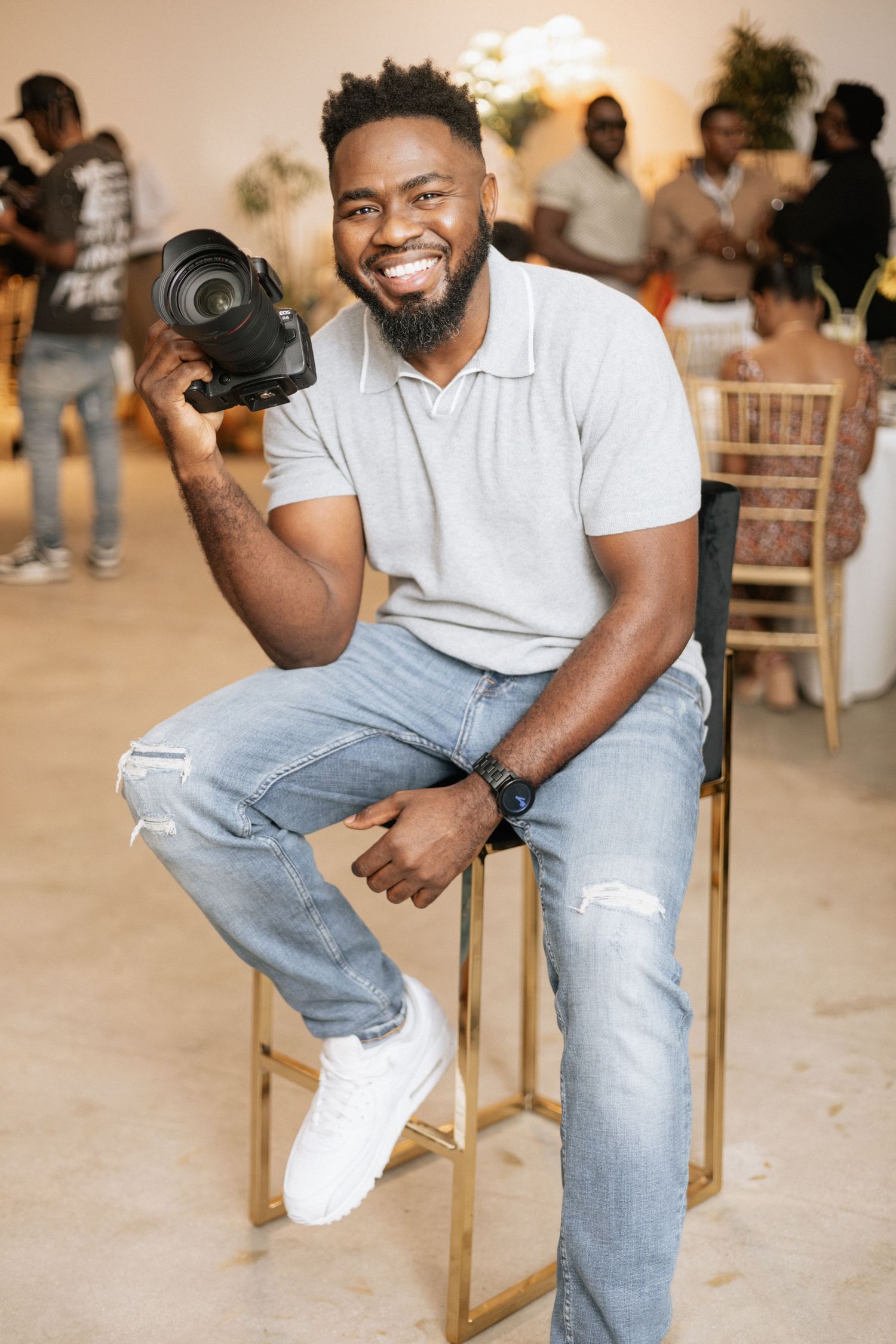 A person in jeans and white polo shirt poses on wooden chair in front of blurred background.