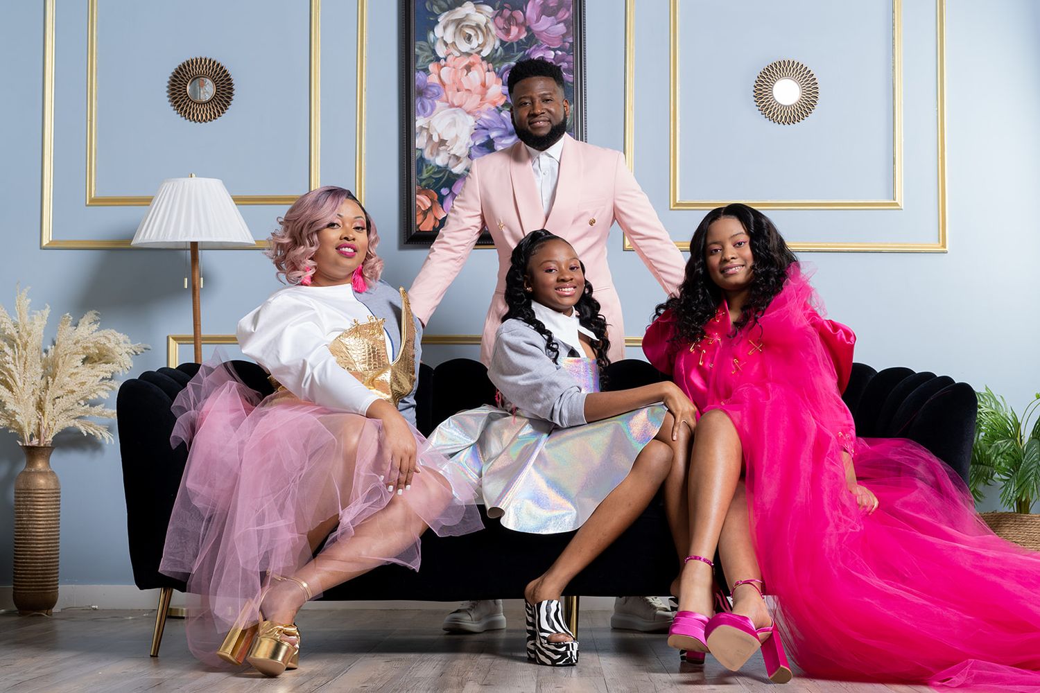 Four people pose together on a couch in coordinated pink and white outfits against a blue wall with elegant decor.