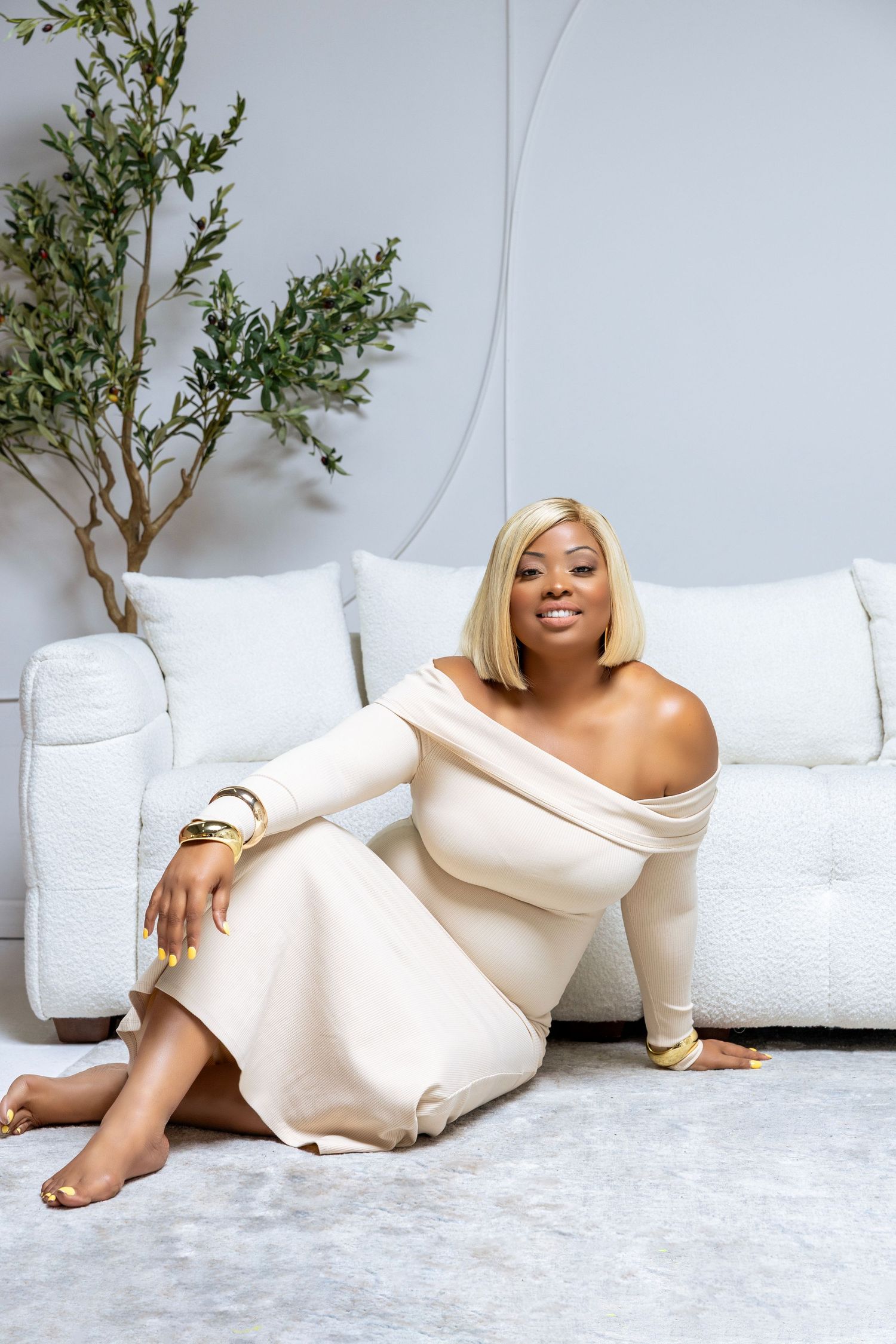 Elegant off-shoulder cream dress on minimalist white couch with small green plant decor in background.