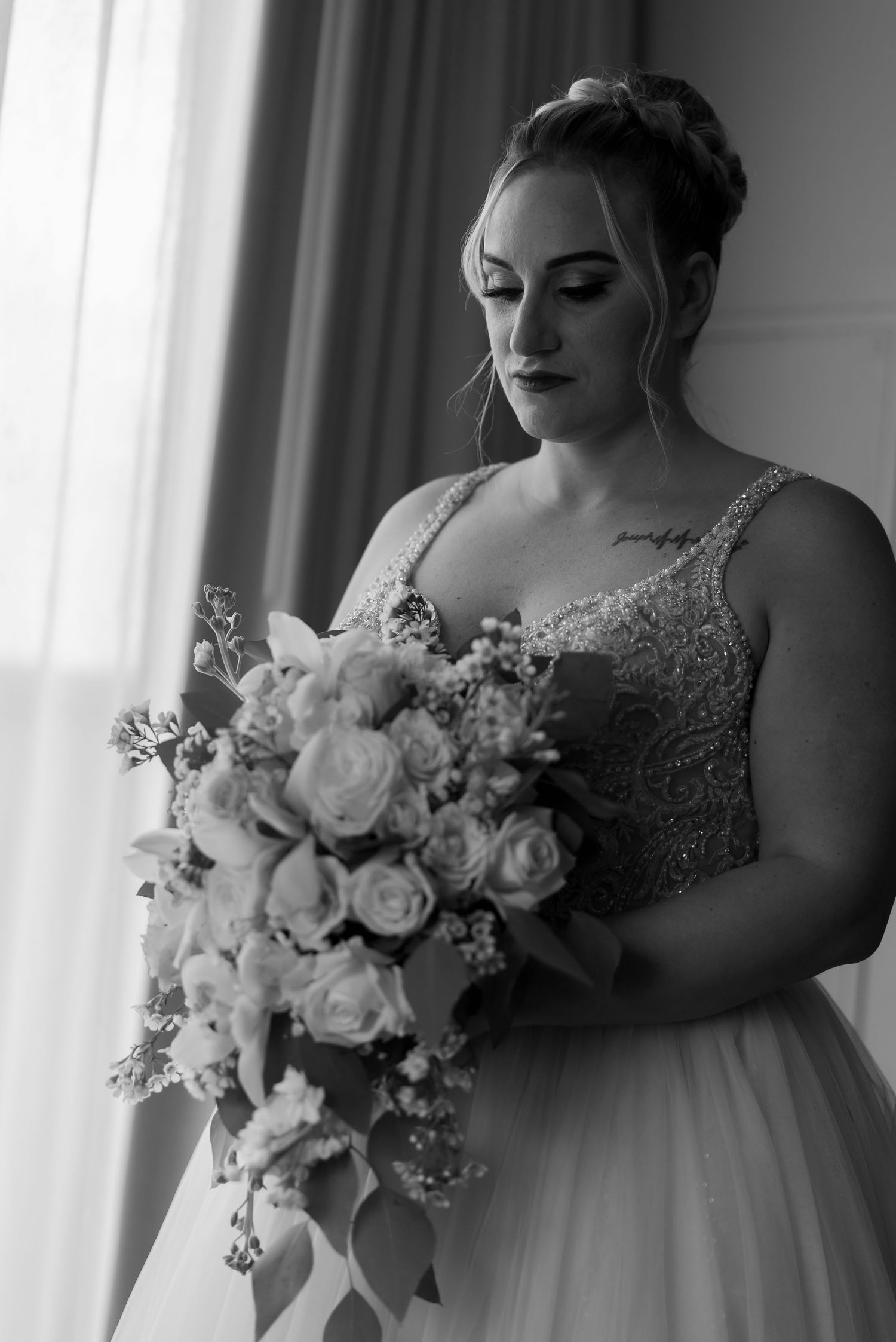 A bride in a flowing ballgown wedding dress holds a romantic bouquet of white roses near a window in black and white.
