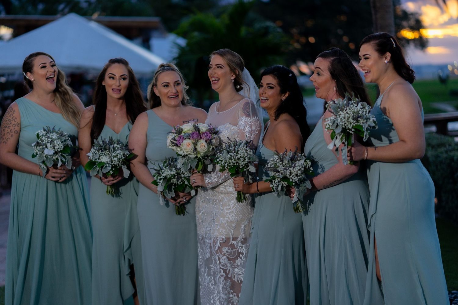 Wedding party in sage green bridesmaid dresses holding white floral bouquets at sunset outdoor ceremony.