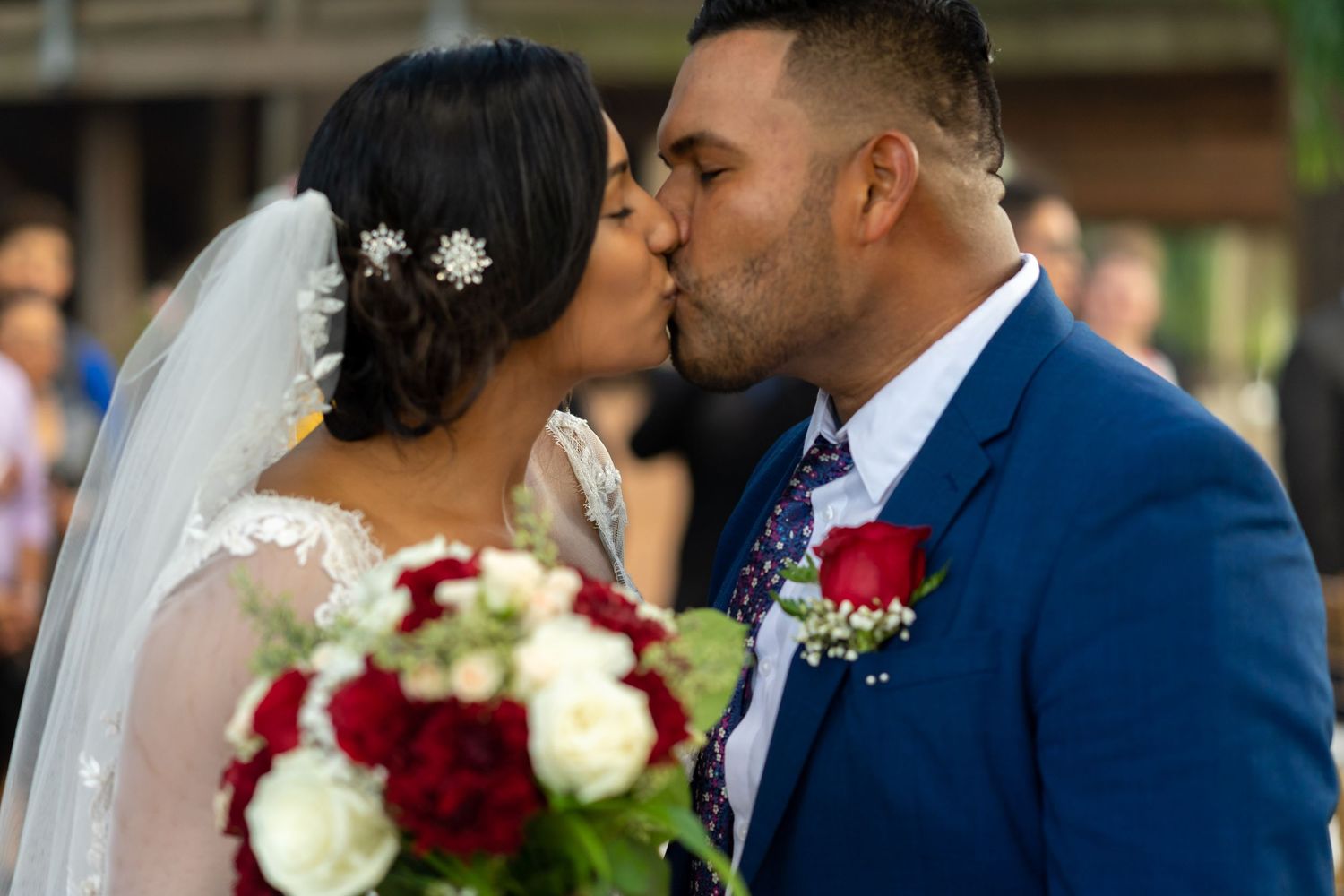 A bride and groom share an intimate kiss during their wedding ceremony with red and white floral decor in the background.