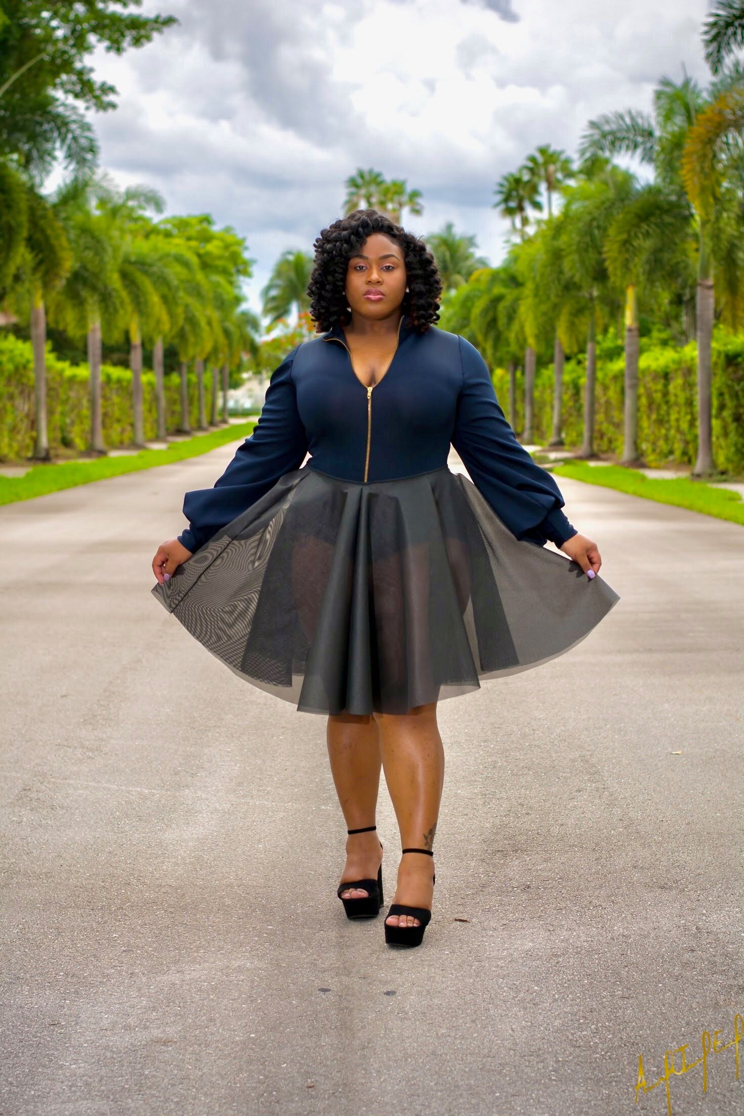 A person in a navy blue top and flared tulle skirt poses confidently on a palm tree-lined path.