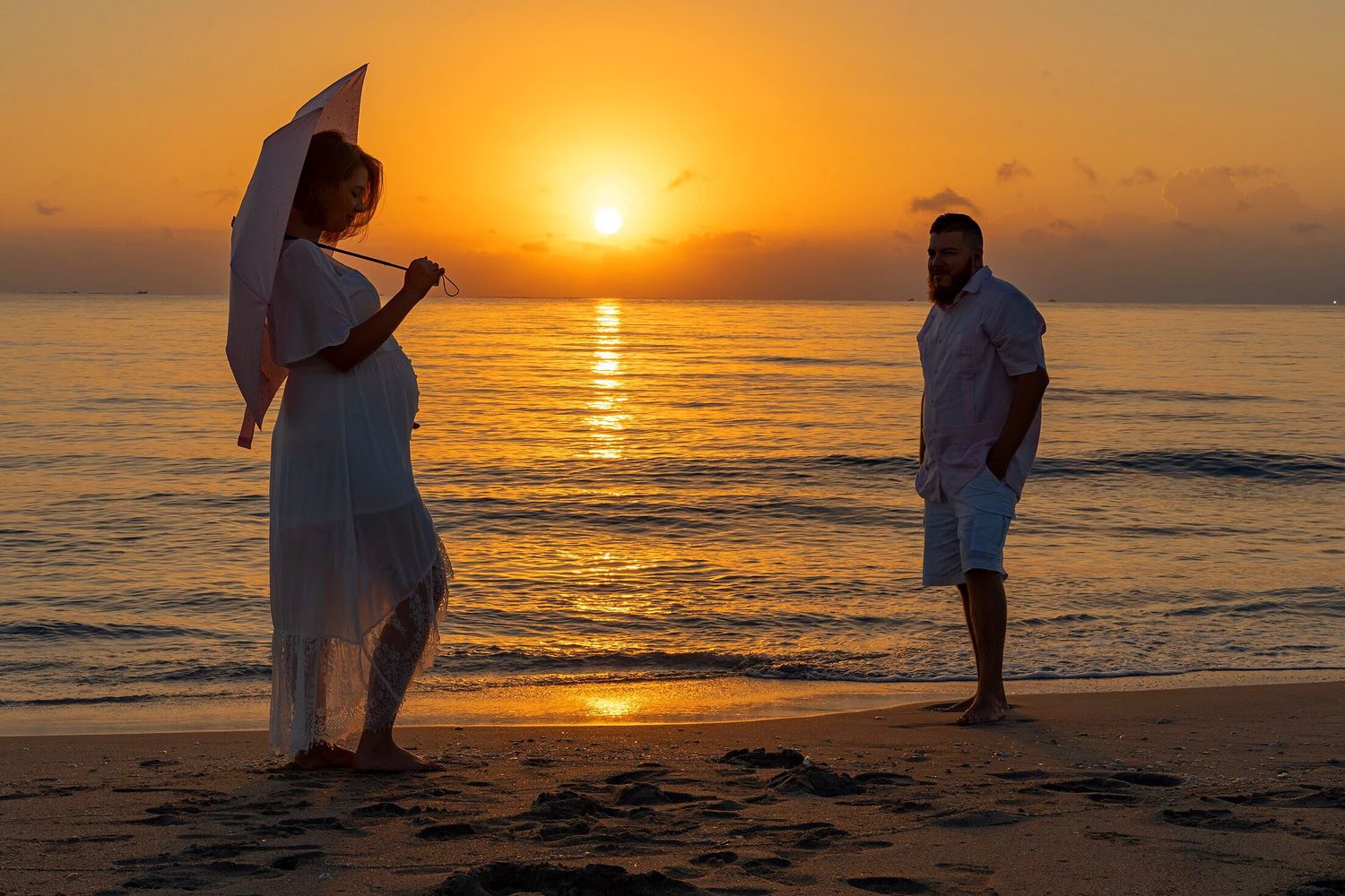 A couple in white attire stands on a beach at sunset with one holding a parasol near the ocean.