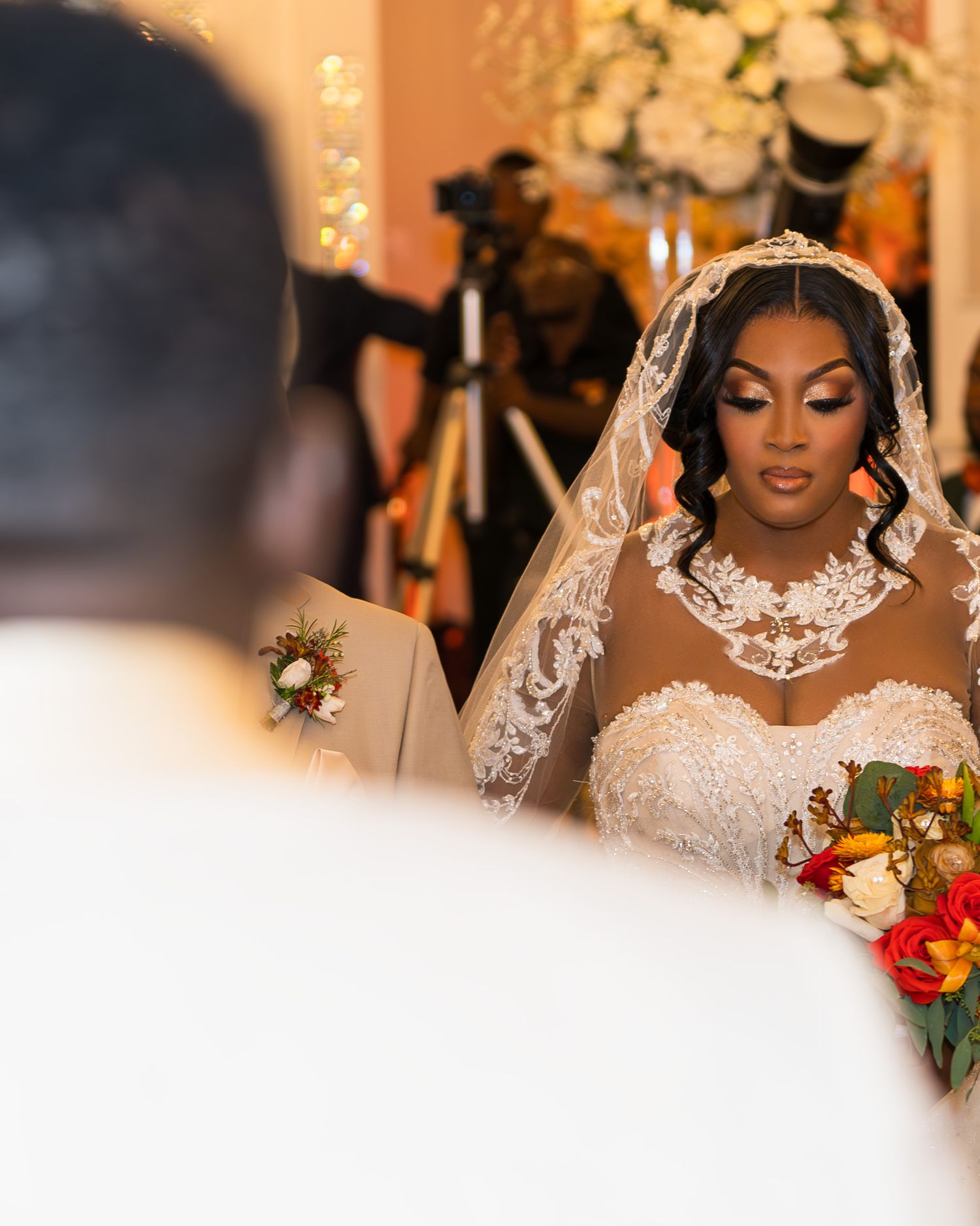 A bride in an elegant white lace veil and strapless gown with sparkling necklace holds a vibrant autumn bouquet during ceremony.