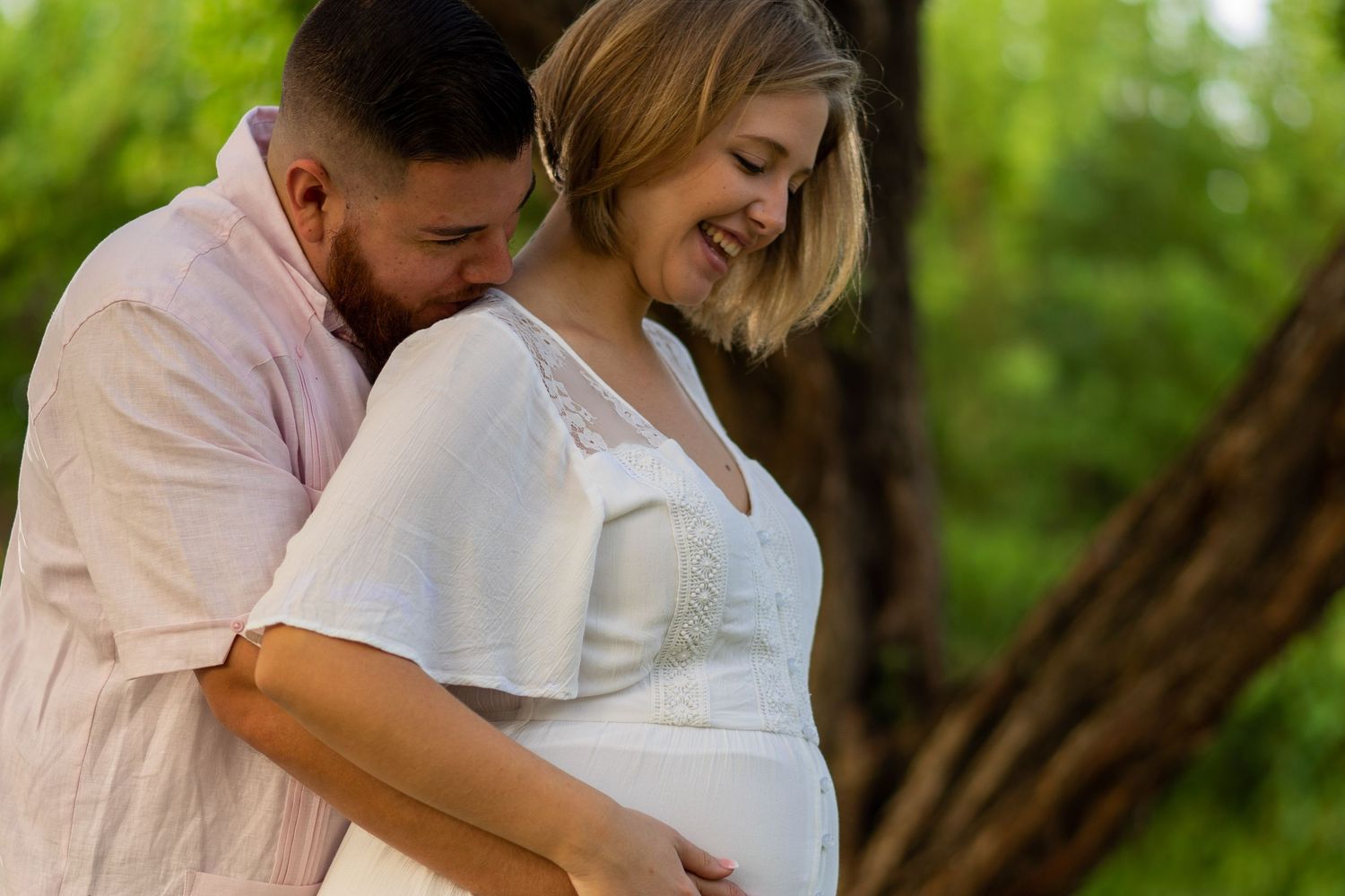 A loving couple embraces outdoors during a maternity photo session with lush greenery in the background.