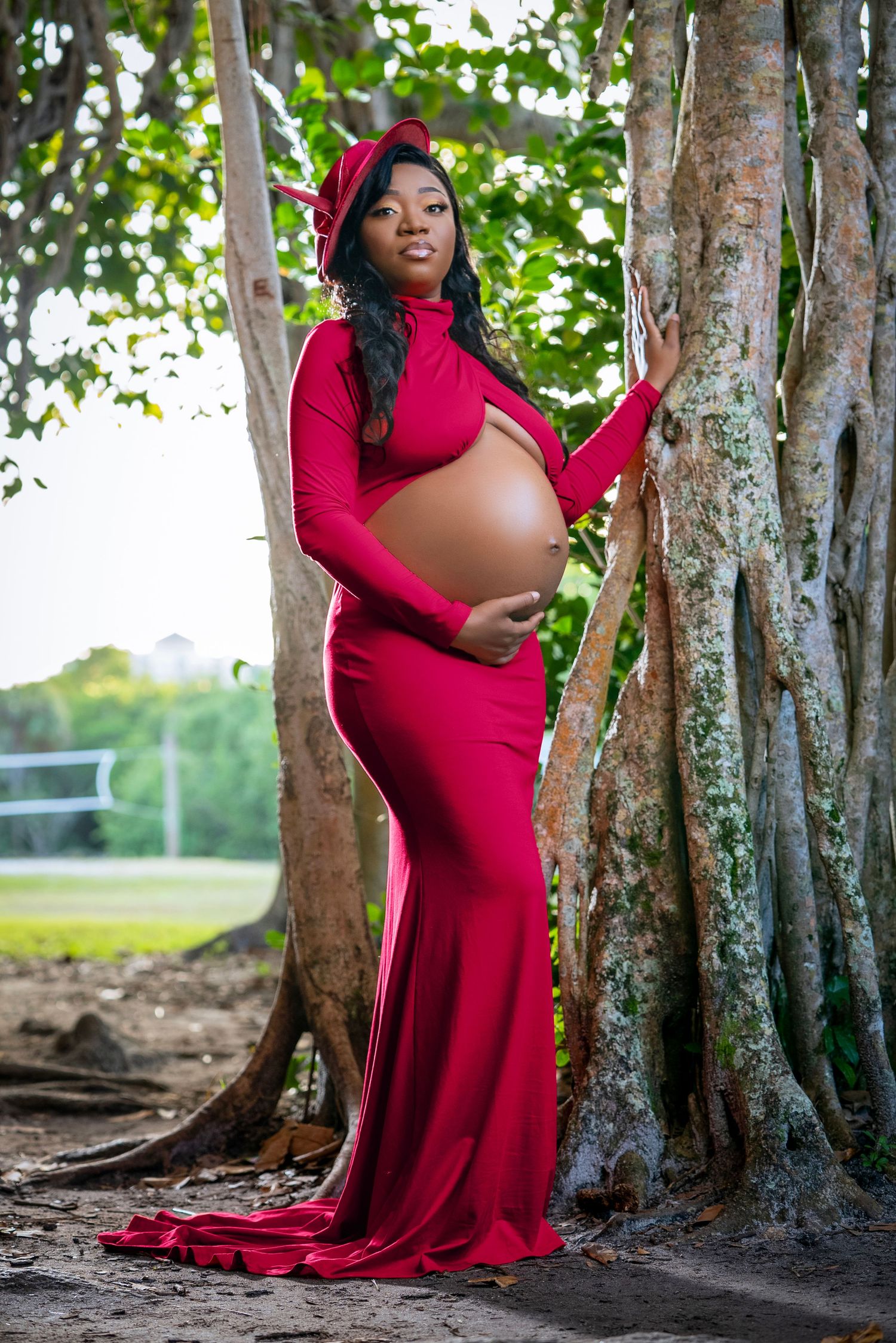 A pregnant figure in a flowing red gown and matching beret poses elegantly against a large tree trunk in a natural setting.